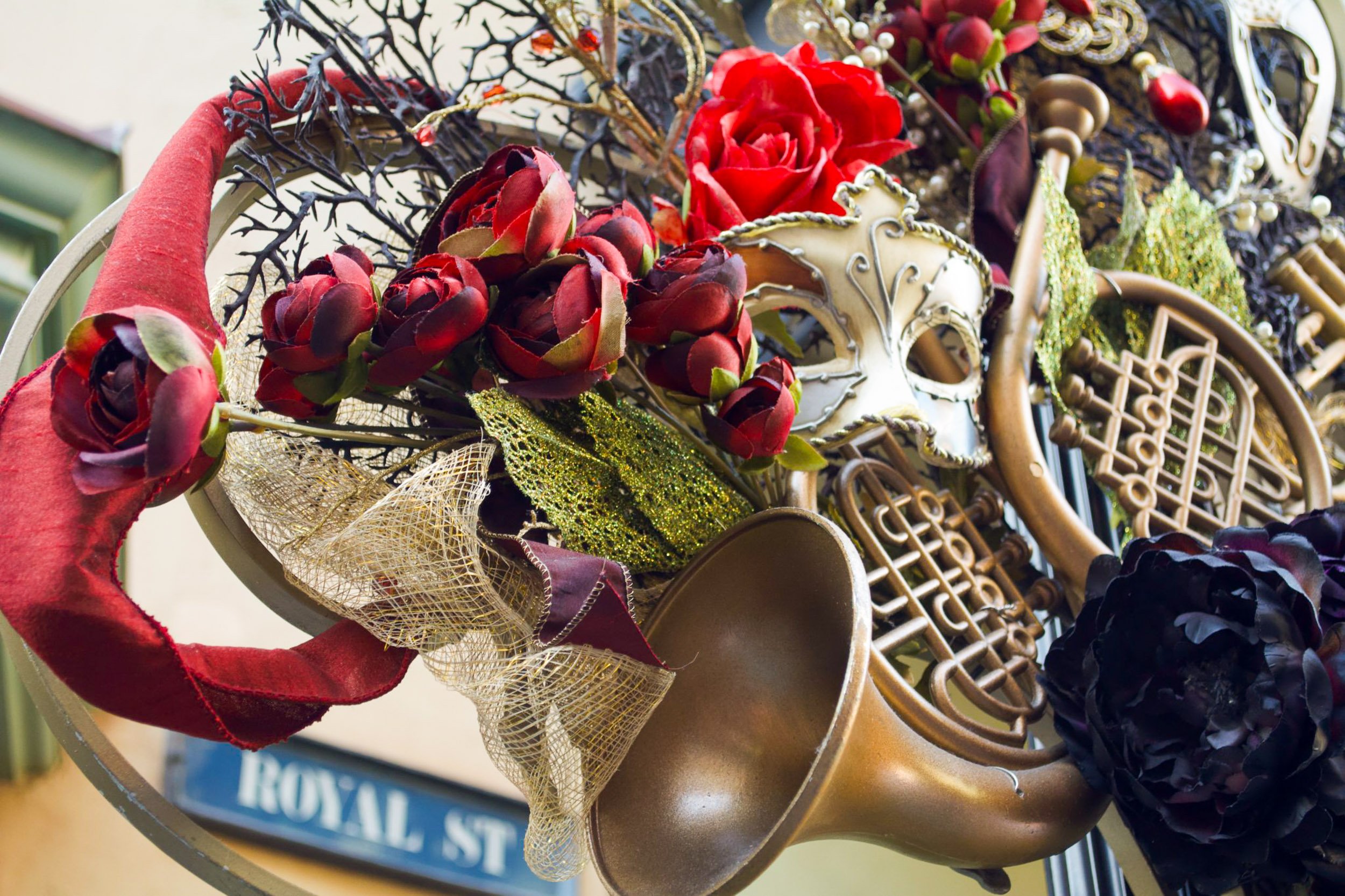 Mardi Gras Melody - A rich, textured close-up of a festive Royal Street sign display adorned with deep red roses, a gold masquerade mask, glittery green leaves, and a vintage brass horn instrument.