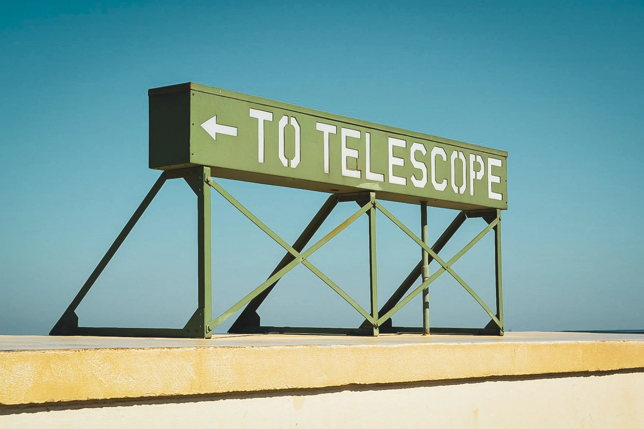 To Telescope - A minimalist, low-angle shot of a vintage green directional sign at the Griffith Observatory, set against a stark, cloudless coastal sky.
