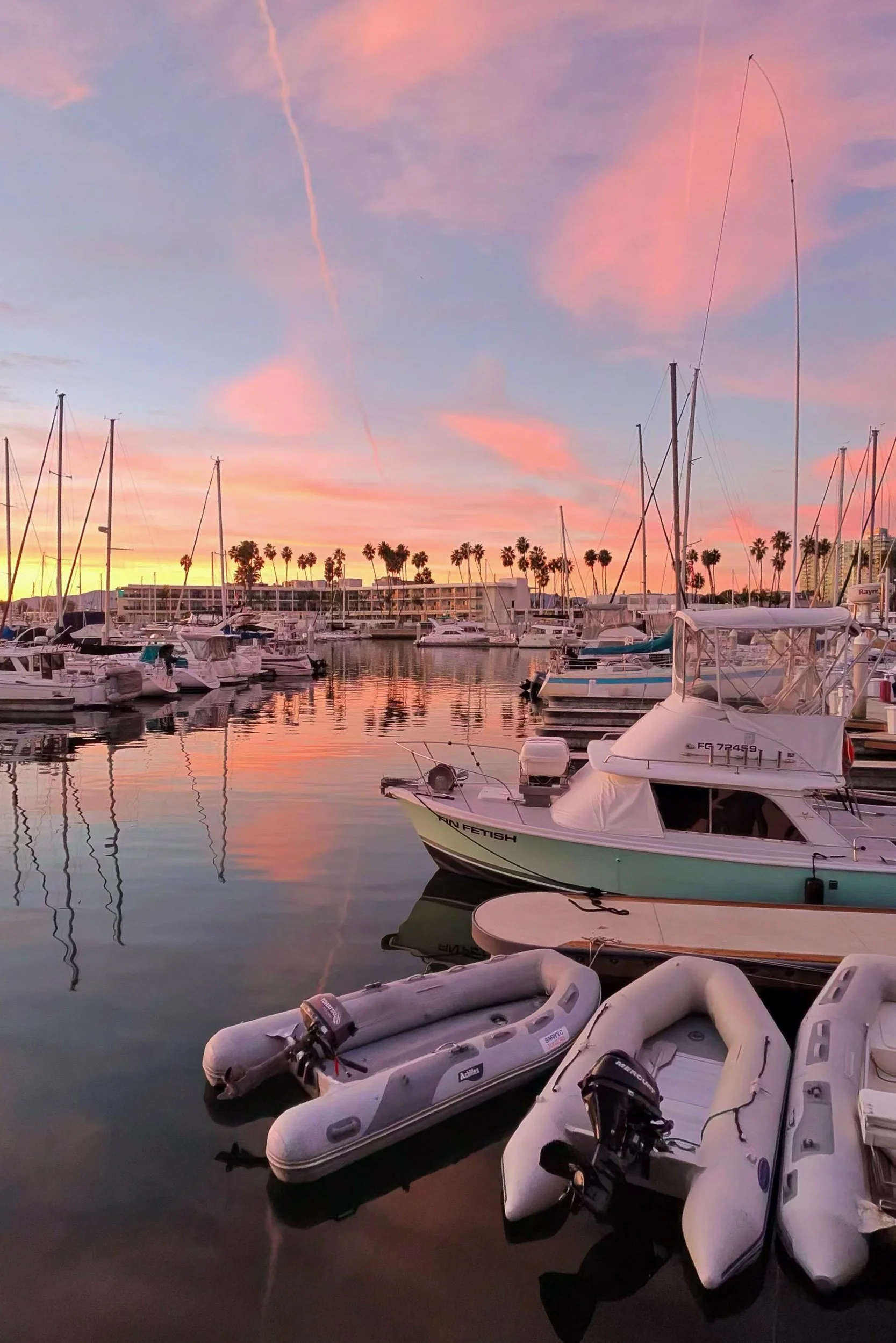 Harbor Glow - A brilliant pink and orange sunset sky reflecting over a calm marina filled with docked boats, yachts, and silhouetted palm trees.