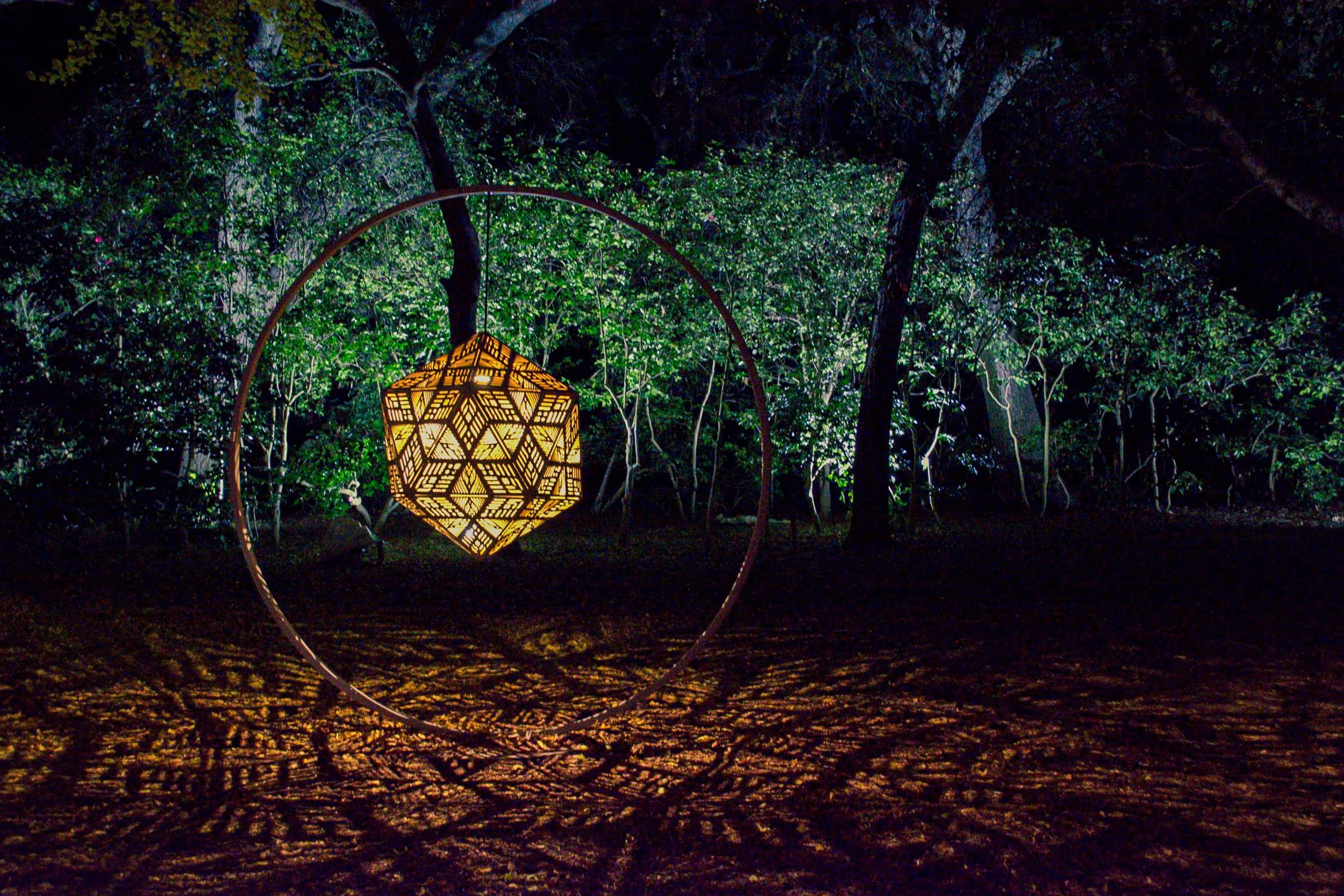 Geometric Glow - A large, intricate geometric lantern suspended in a dark forest, casting warm, patterned shadows across the ground at night.