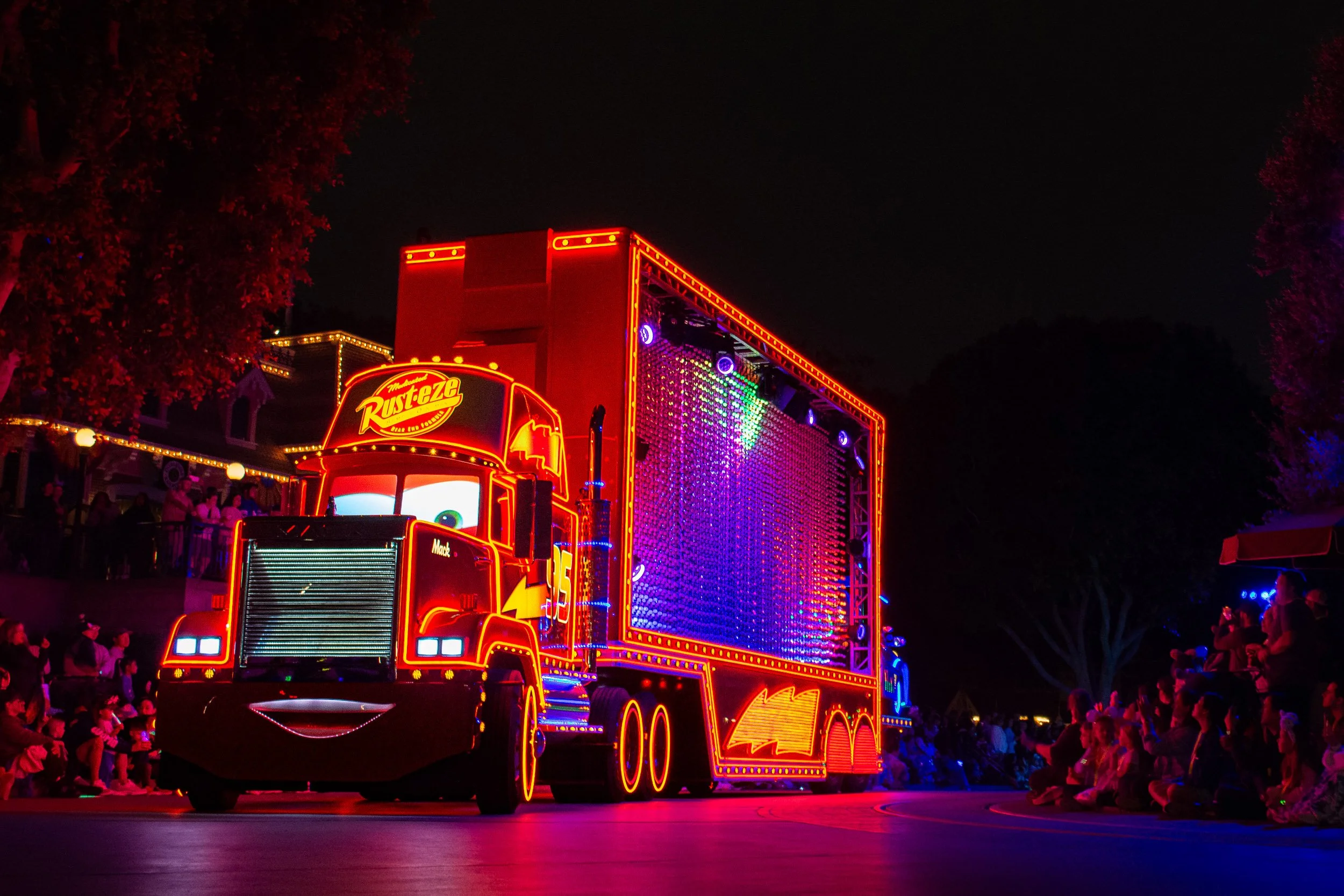 Neon Haul - A vibrant nighttime parade photograph of the iconic Mack truck float completely outlined in brilliant red and orange neon lights, moving past a dark crowd with its trailer side panel glowing in intense purple and blue light.