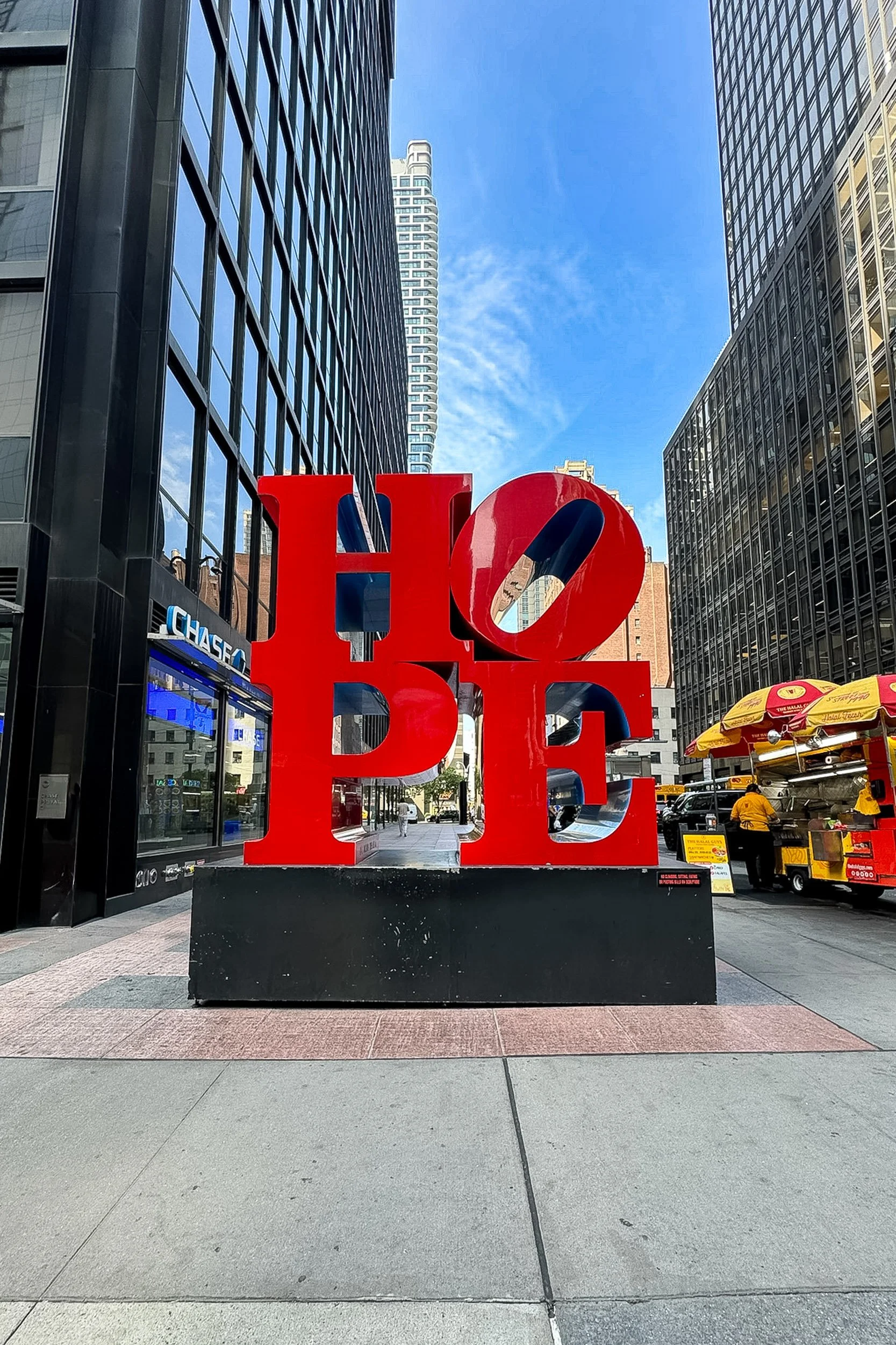 Urban Hope - A vibrant, large red HOPE sculpture standing boldly on a city sidewalk framed by towering glass office skyscrapers and a bright blue sky.