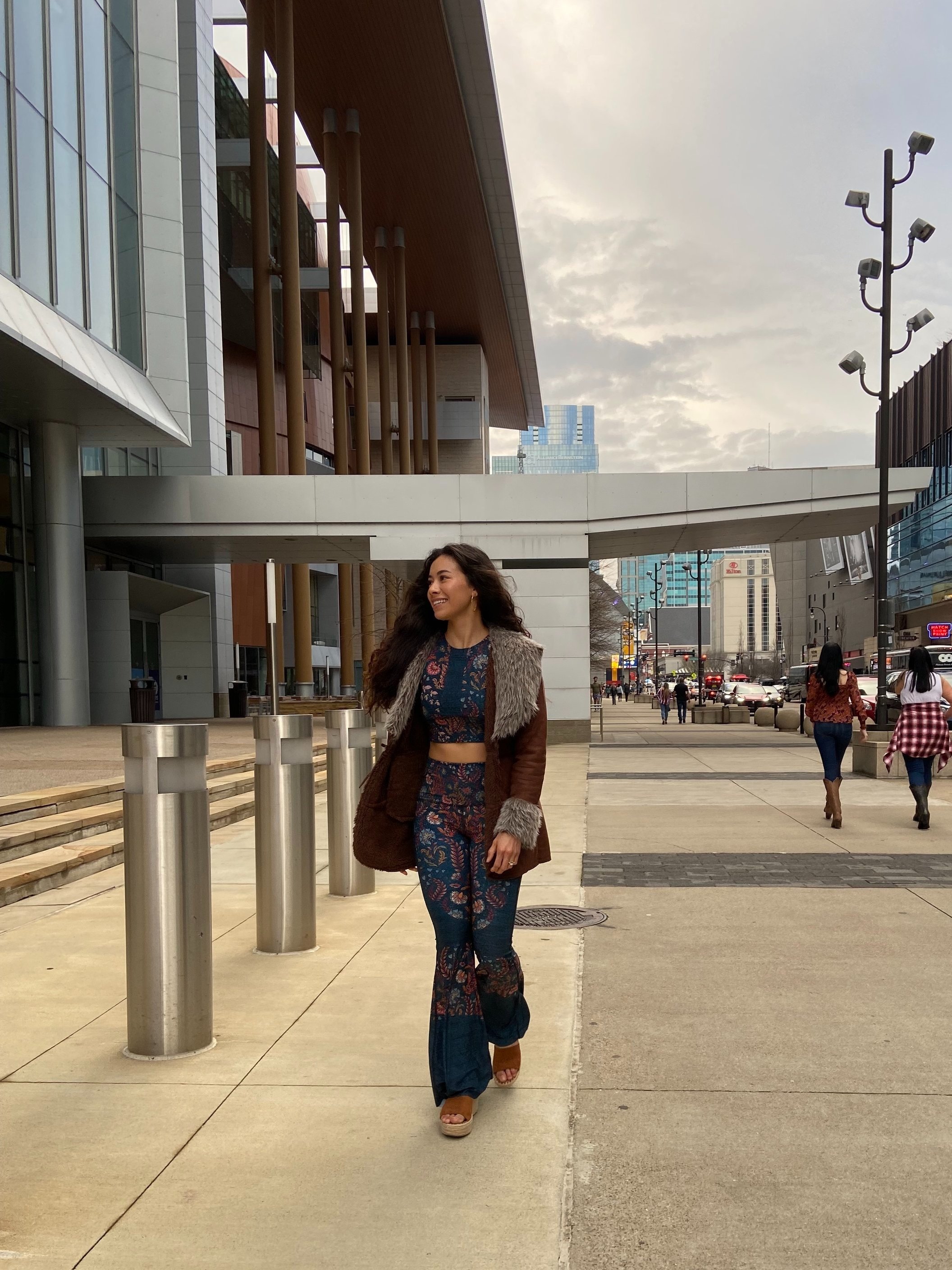 A woman with long curly hair wearing a floral outfit, a brown fur-lined coat, and platform sandals walking in an urban area with modern buildings and other pedestrians.