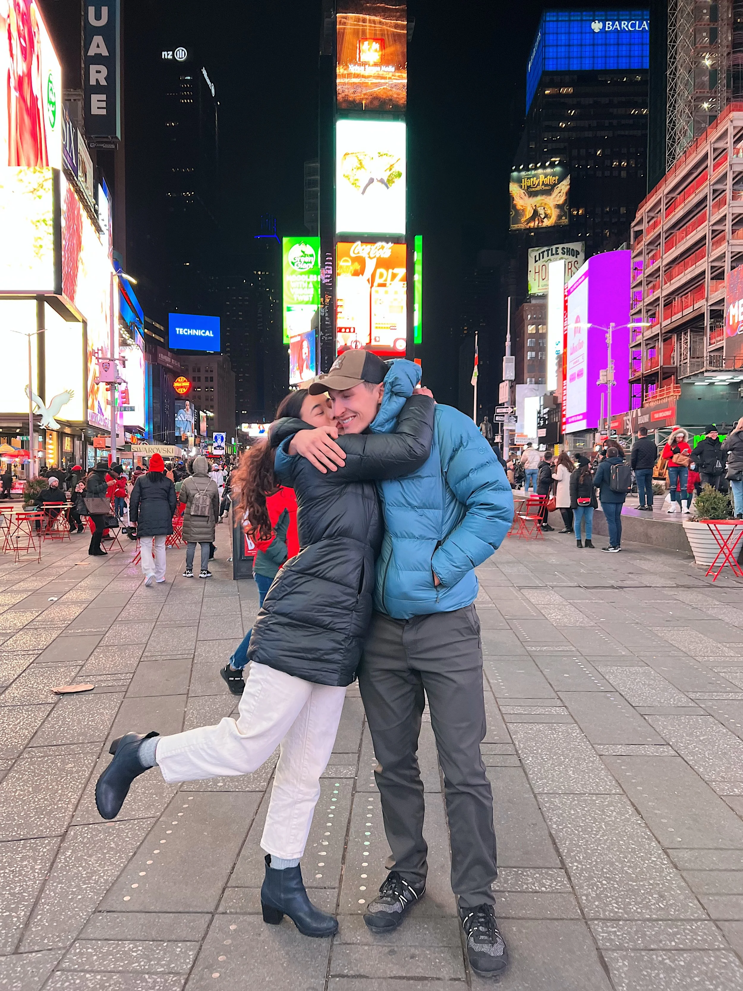 Two people hugging in Times Square at night, one person lifting the other. Bright lights and billboards surround them, with crowds of people in the background.