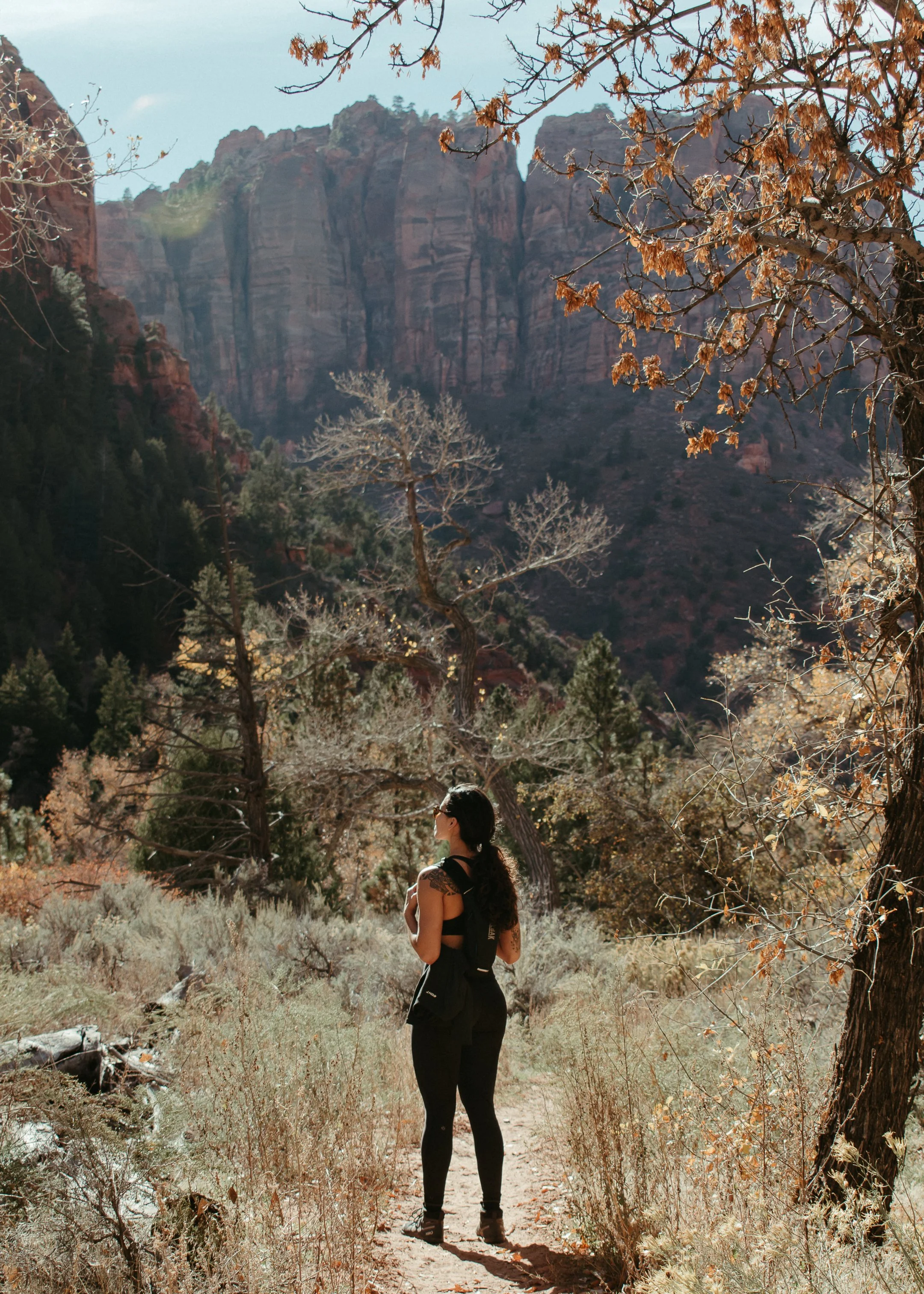 A woman with long dark hair wearing sunglasses, black athletic clothing, and hiking boots stands on a trail surrounded by dry grass and bushes, with trees and large red rock formations in the background, suggesting a desert or canyon landscape.