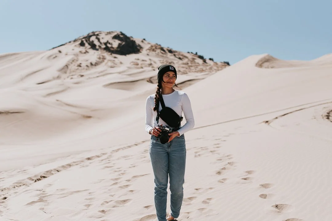 A woman standing on sand dunes in a desert with a camera in her hands, wearing a black beanie, a white long-sleeve shirt, and jeans.
