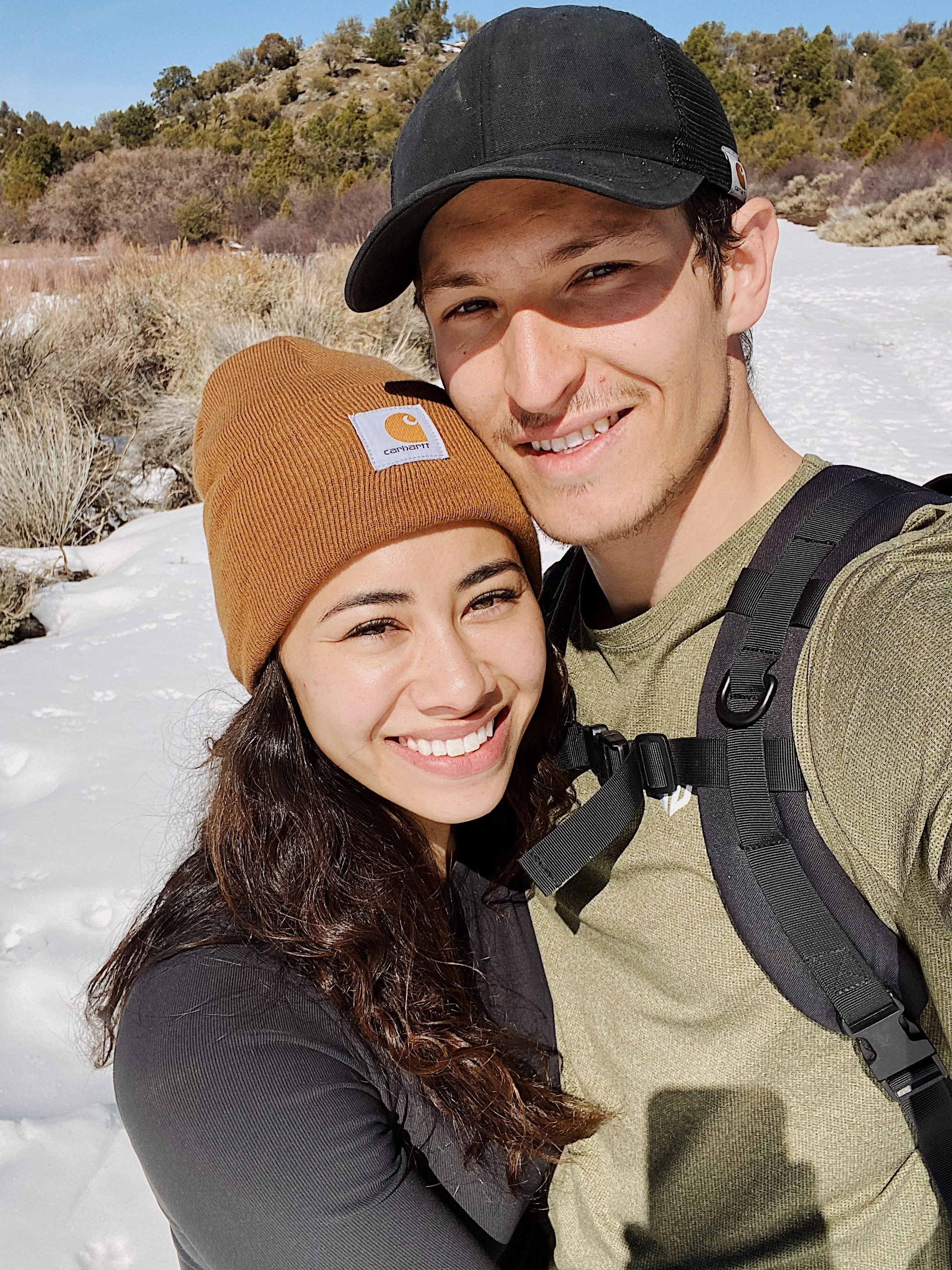 A young couple takes a selfie outdoors in a snowy landscape with bushes and trees in the background. The woman has long dark hair and wears a brown beanie with a Carhartt logo. The man has short dark hair and wears a black cap and a backpack. They ar