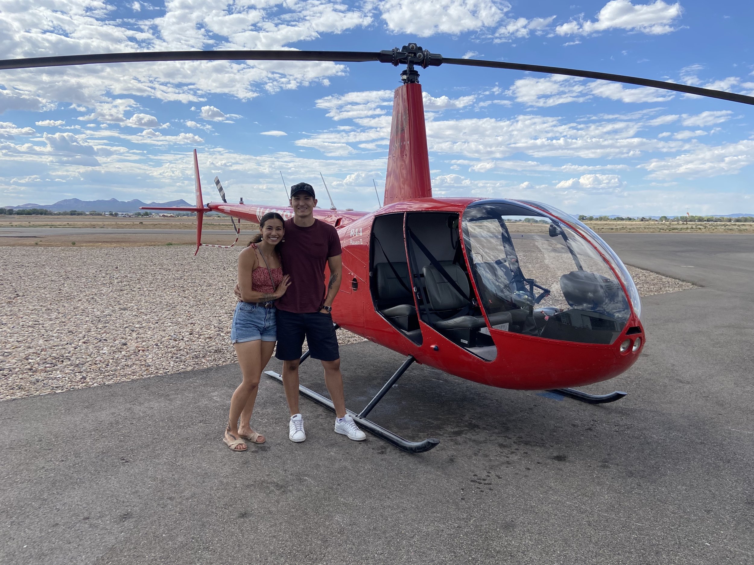 Two smiling people, a woman and a man, standing side by side in front of a Southern Utah University helicopter on a runway, with mountainous terrain and a partly cloudy sky in the background.