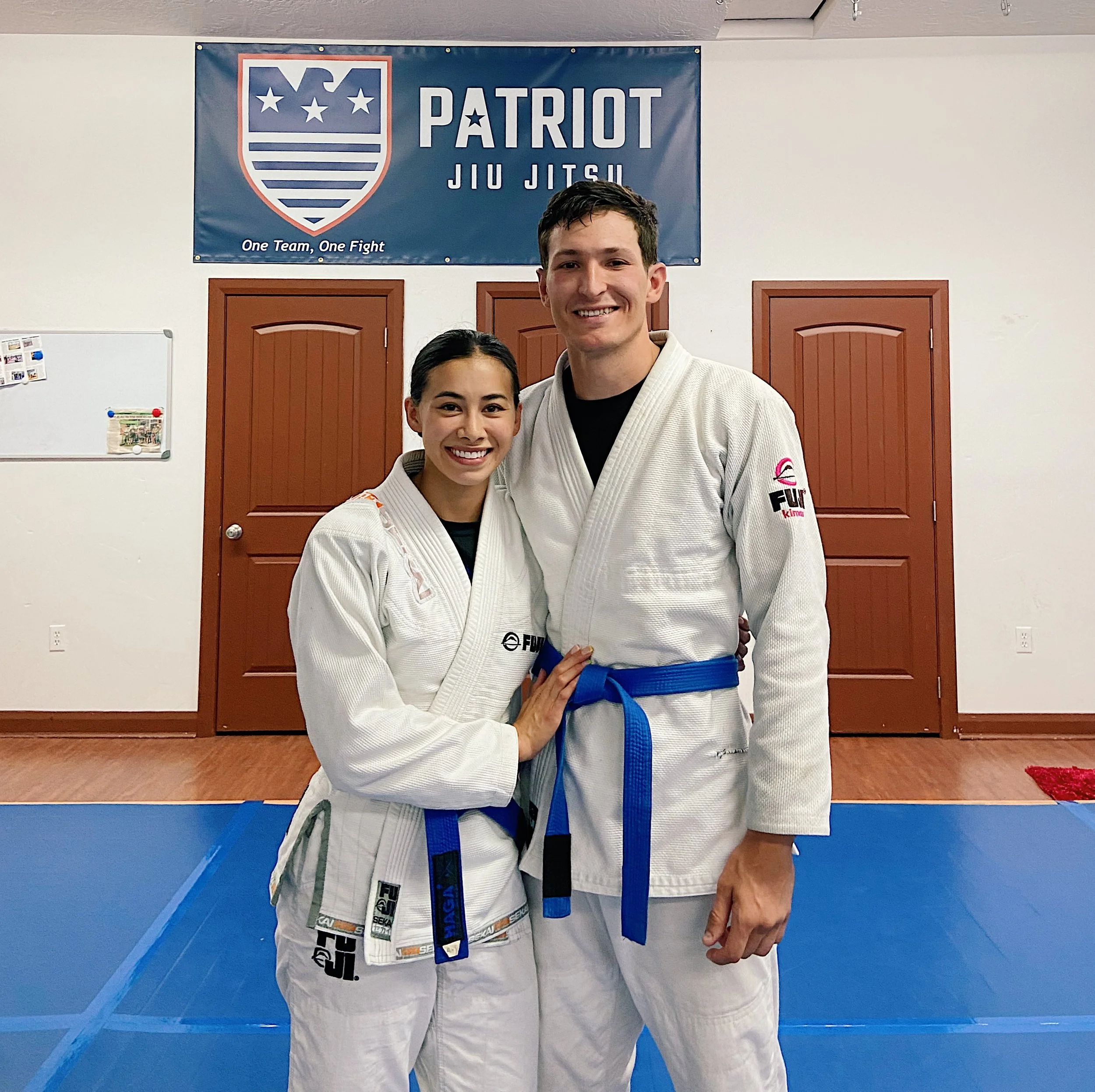 Two individuals in martial arts uniforms posing for a photo in a dojo, with a Patriot Jiu Jitsu banner in the background.