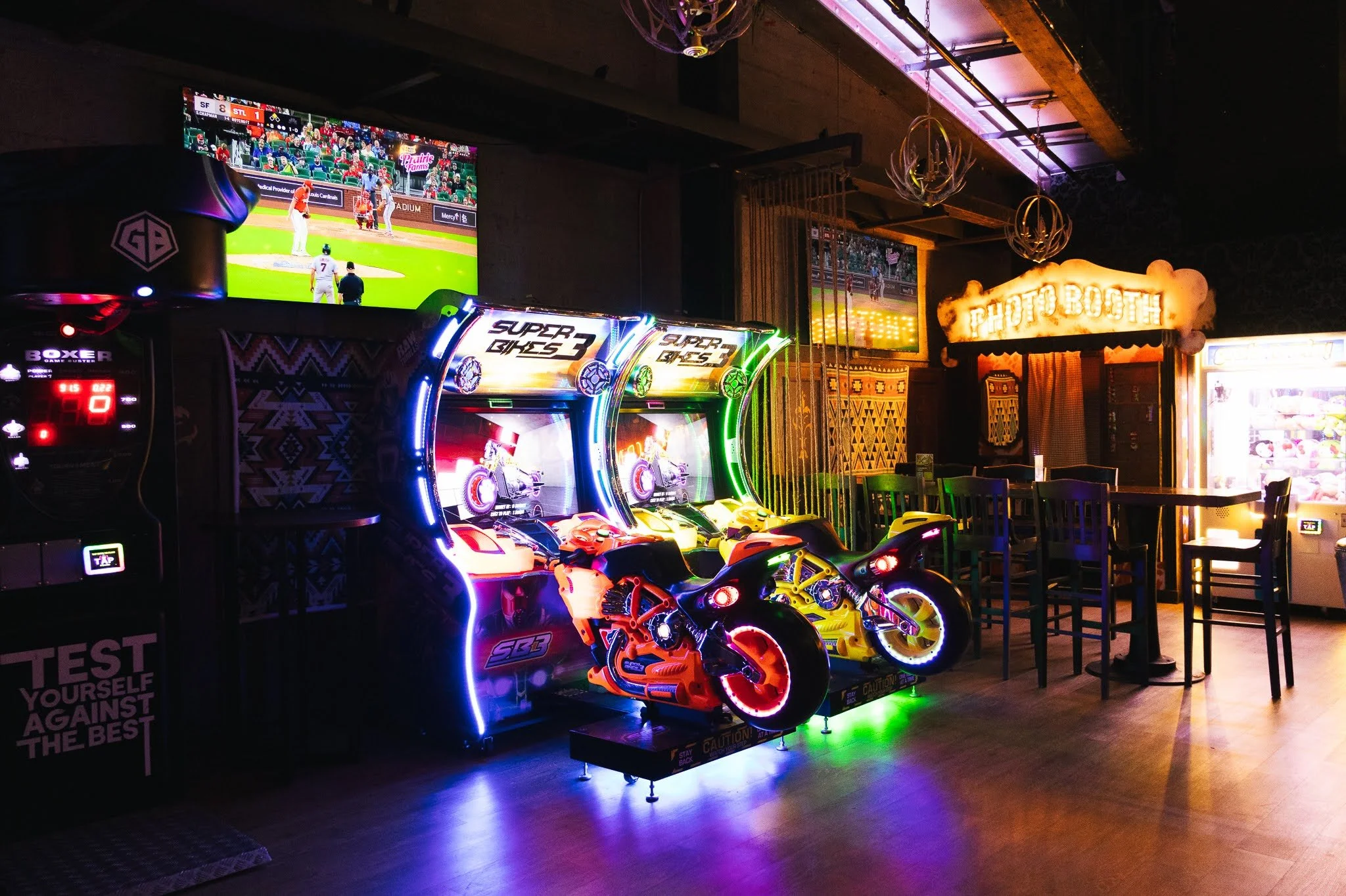 Indoor arcade featuring motorcycle racing games with illuminated neon signs, a photo booth, a claw machine, and multiple television screens displaying sports.