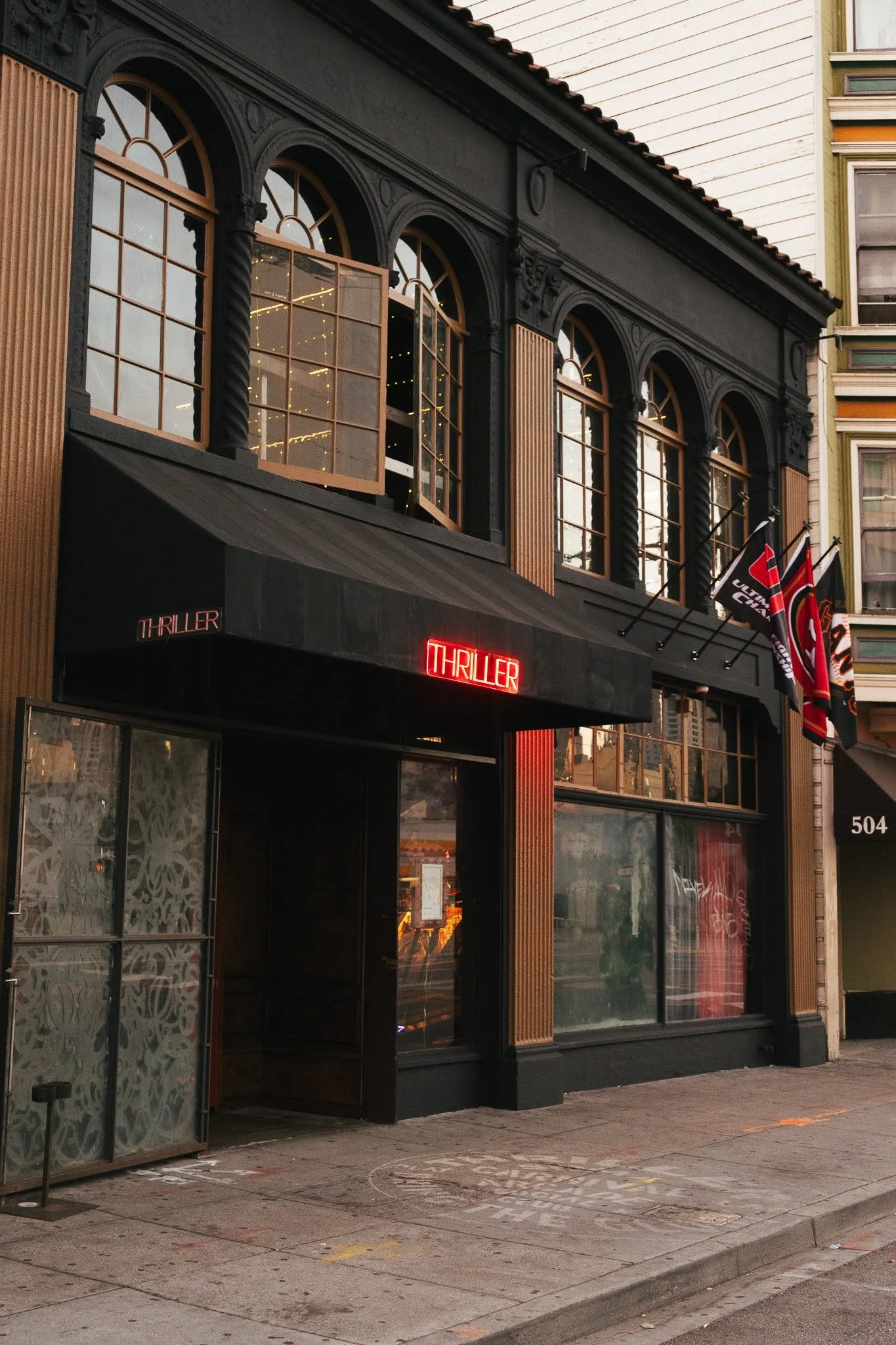 The exterior of a building with black and wooden architectural details, several illuminated 'Thriller' signs, and flags hanging outside, with a city street in front.