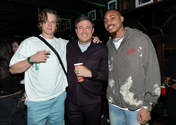 Three men smiling and posing together at a social event. The man in the middle is holding a red cup, the man on the left has a green wristband, and the man on the right is wearing a distressed gray hoodie. They are indoors with a dark background and 