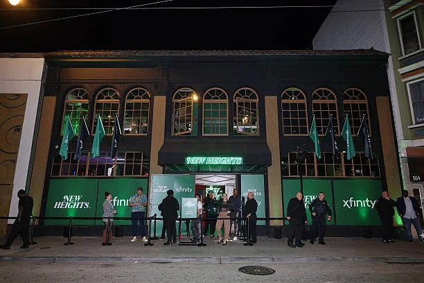 People are standing outside a building with green signage that reads "New Heights" and "Xfinity." There are multiple flags on poles and a line of people waiting outside, some wearing masks.