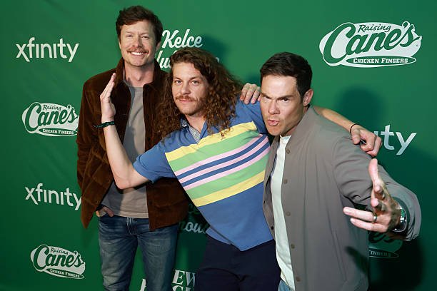 Three men posing in front of a green background with Raising Cane's logo, Xfinity logo, and other branding. The man on the left has a beard and curly hair, wearing a brown jacket and smiling. The man in the middle has long curly hair, wearing a strip