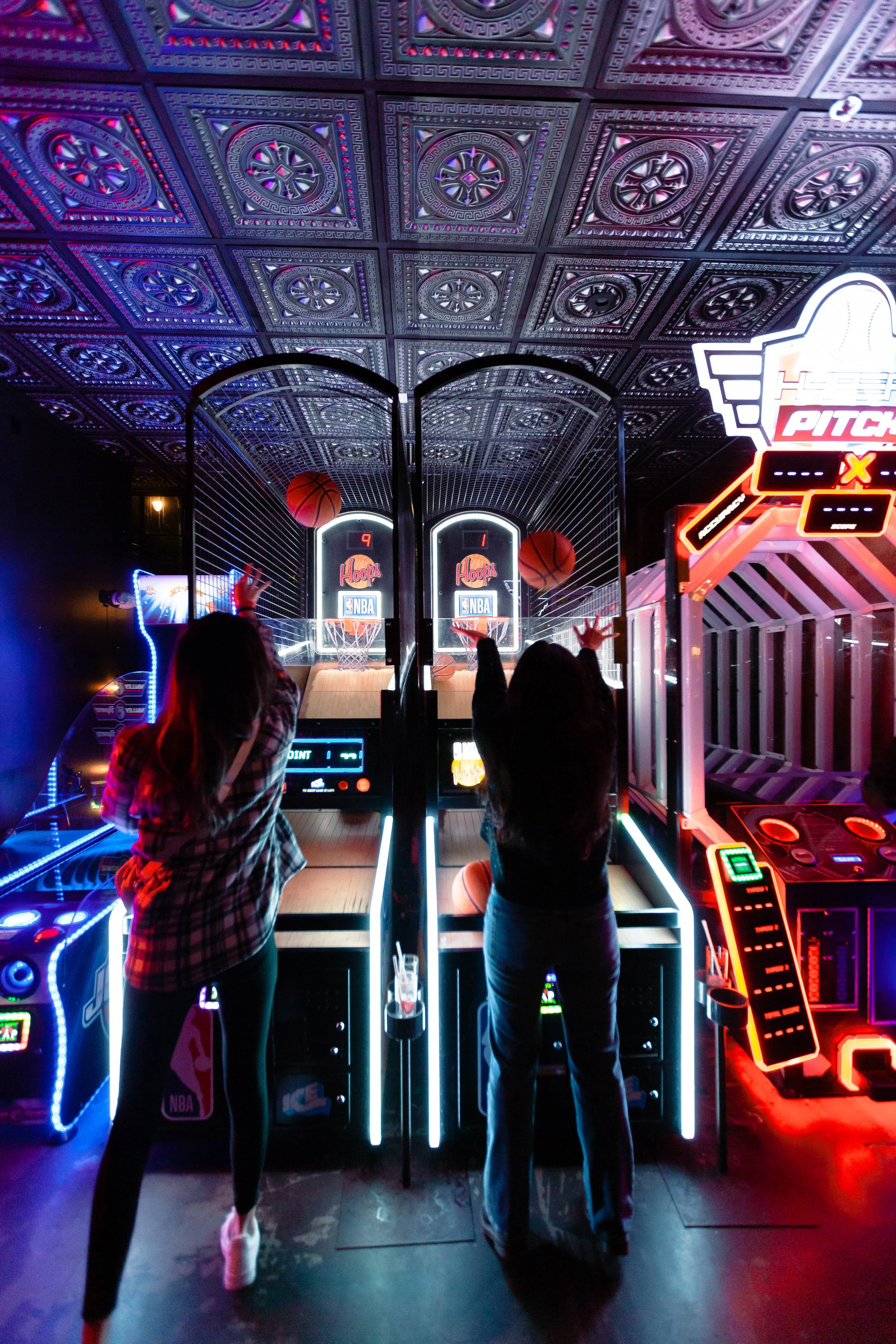 Two women are playing basketball arcade game in an indoor arcade, with neon lights and a decorative ceiling above.