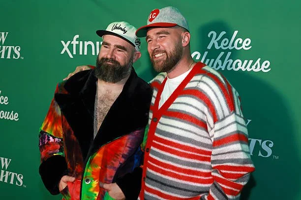 Two men smiling at an event with a green backdrop that has logos and text, including 'Xfinity' and 'Kelce Clubhouse'. One man is wearing a colorful jacket and a white cap with black lettering, while the other is wearing a red, white, and gray striped sweater and a gray cap with a red 'KC' logo.