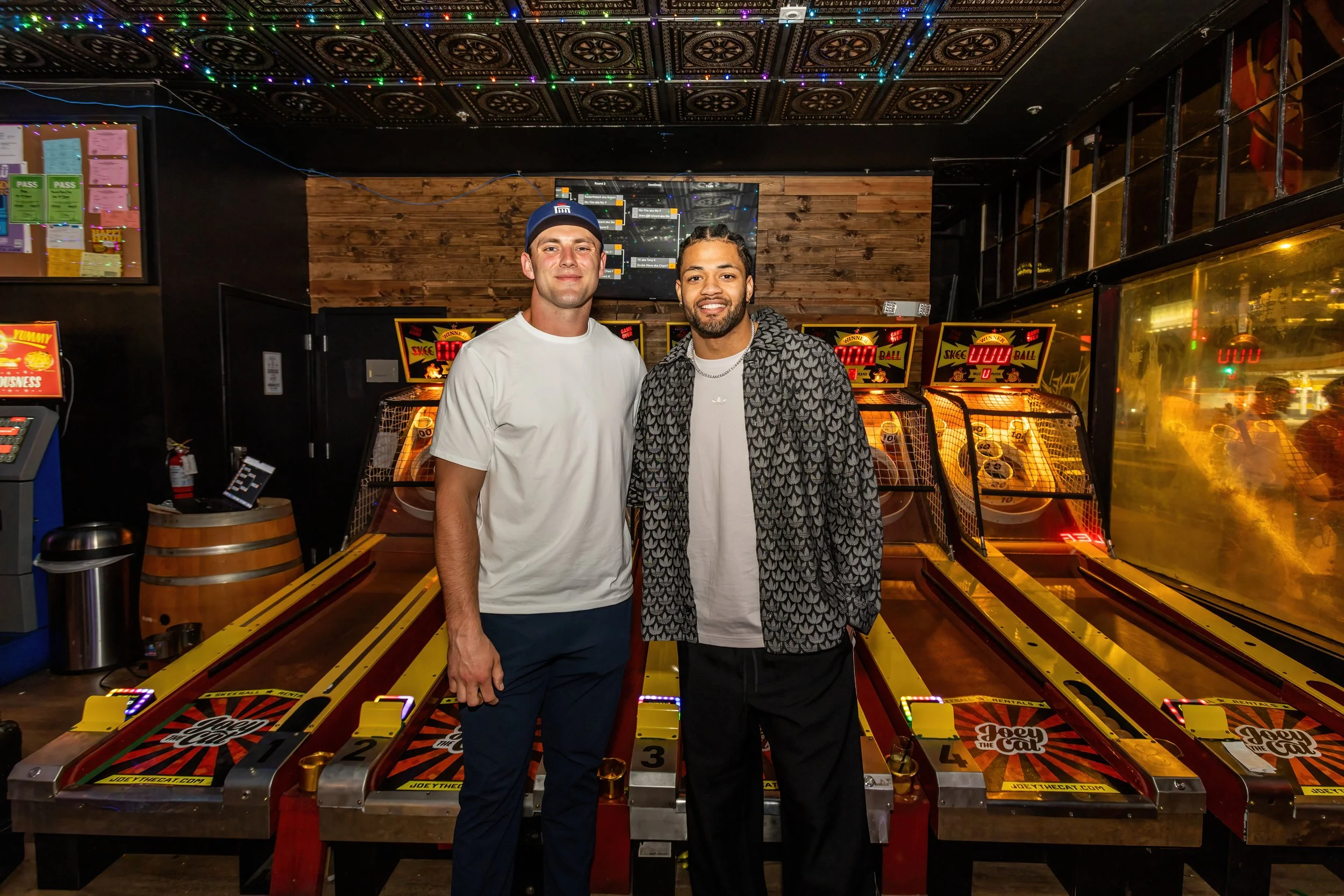 Two men standing together inside an arcade, surrounded by skee-ball machines, smiling at the camera. The arcade has dark walls, a wooden accent wall behind them, colorful string lights on the ceiling, and a window reflecting outside lights.