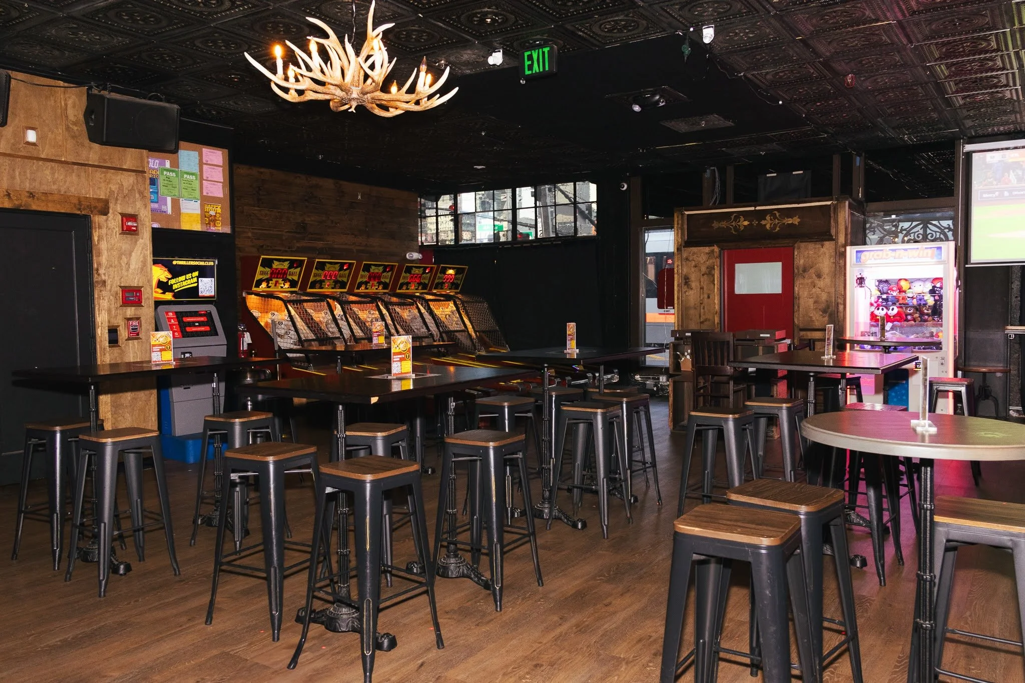 An empty game bar or arcade with high tables, bar stools, cash kiosk, an arcade game machine, a vending machine, a TV showing a sports game, and a chandelier in the ceiling.