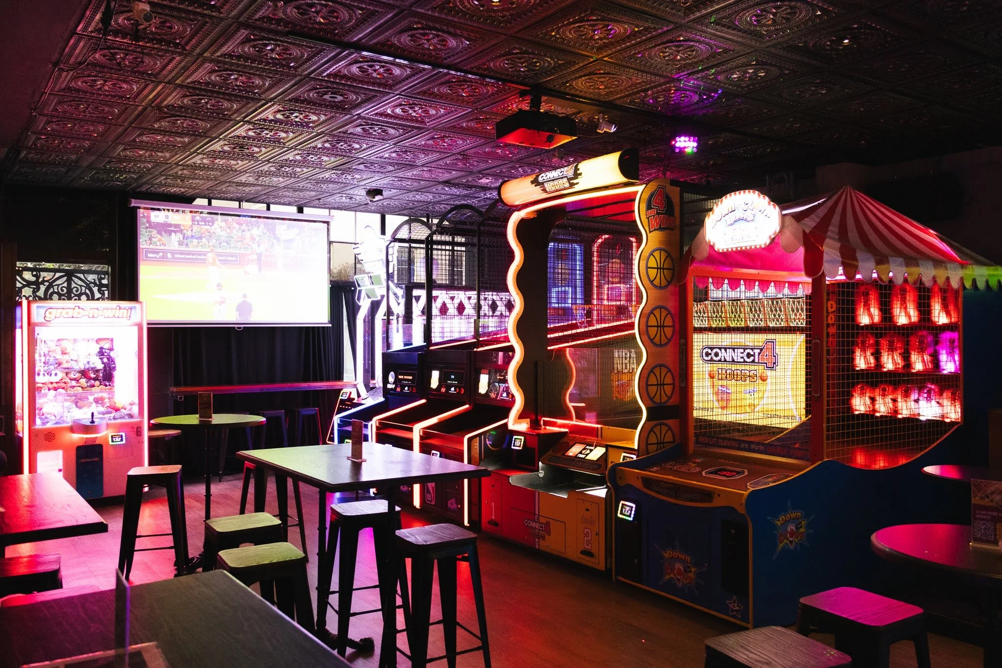 An indoor arcade with various video game machines, a large screen displaying a sports game, and tables with stools. Neon lights and decorative ceiling tiles create a vibrant atmosphere.