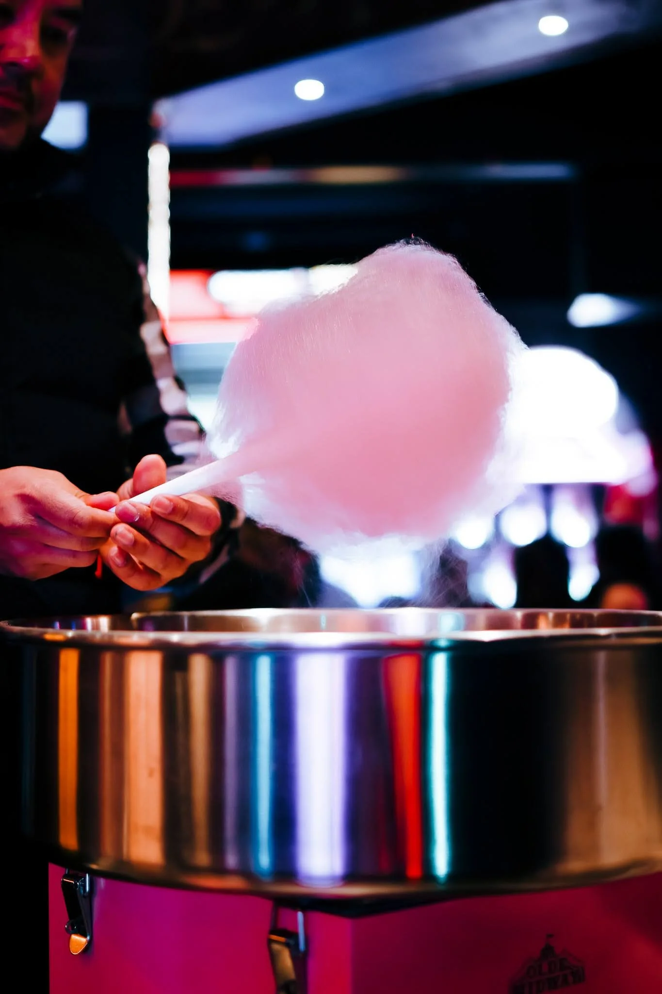 A person is holding pink cotton candy over a shiny metal container, with a dark background illuminated by colorful lights, suggesting an indoor setting like a carnival or entertainment venue.