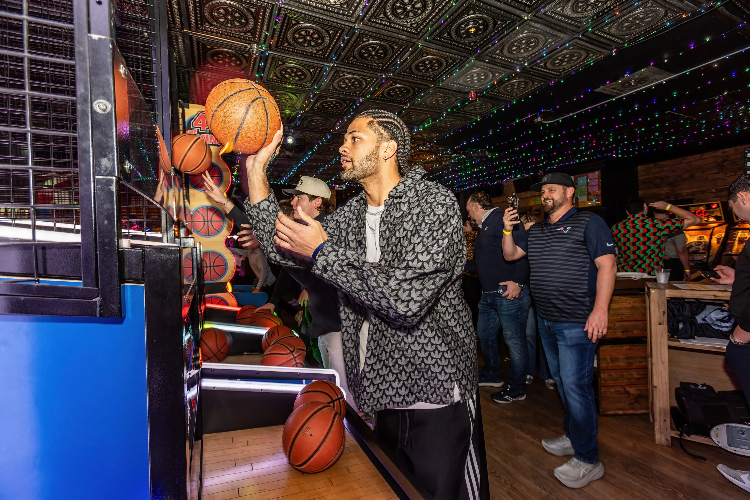 A person with braided hair and a patterned jacket playing basketball arcade game at an indoor amusement center, with other people in the background, colorful string lights overhead, and a wood-paneled wall.