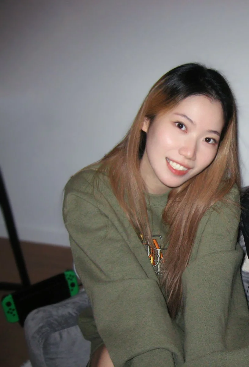 A young woman with long, straight, light brown hair, smiling at the camera. She is wearing a green sweatshirt and is seated indoors, with a partial view of a gaming console and a gray blanket nearby.