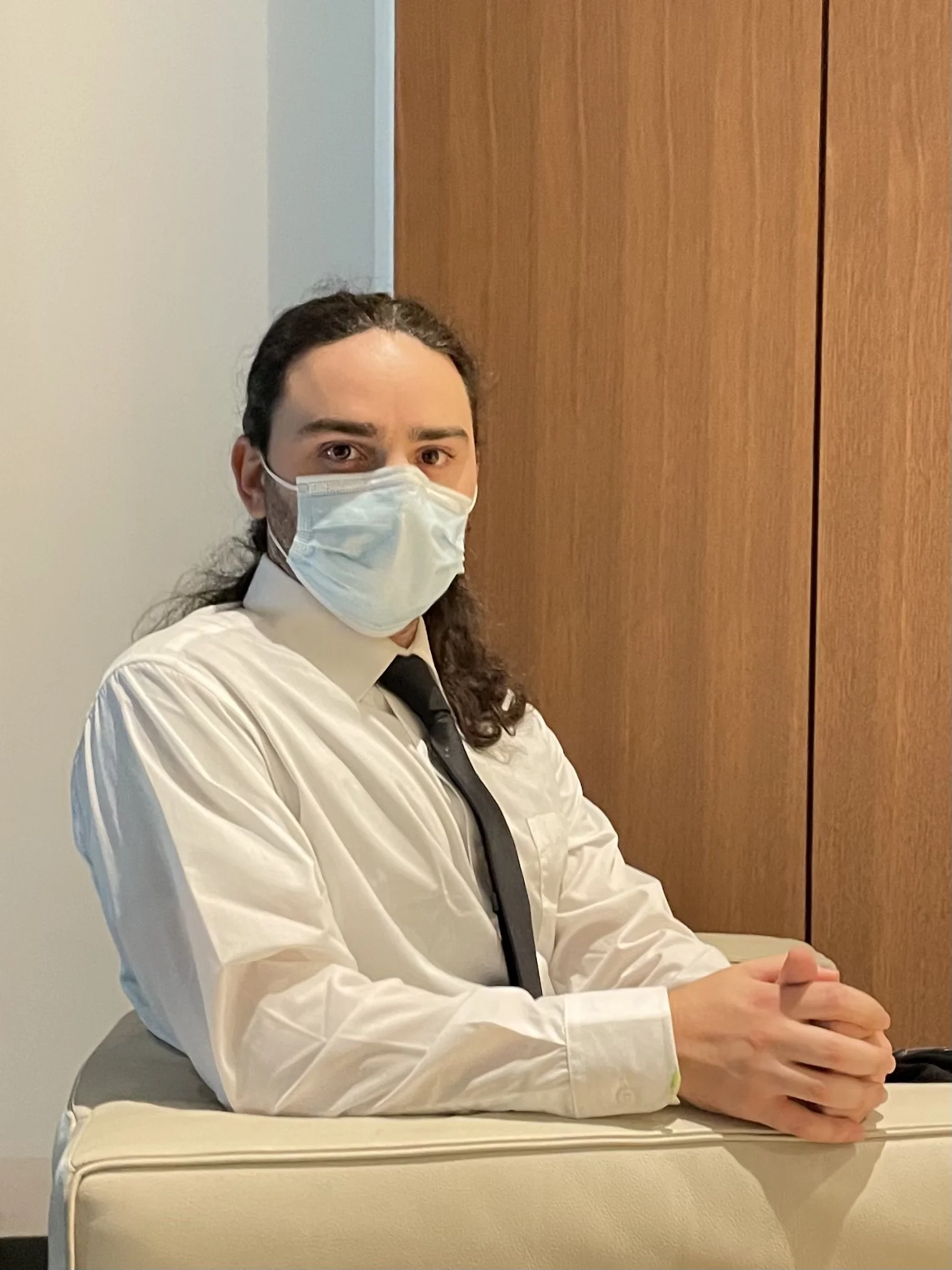 A man with long dark hair wearing a face mask, white shirt, and black tie is sitting at a table in front of a wooden wall.