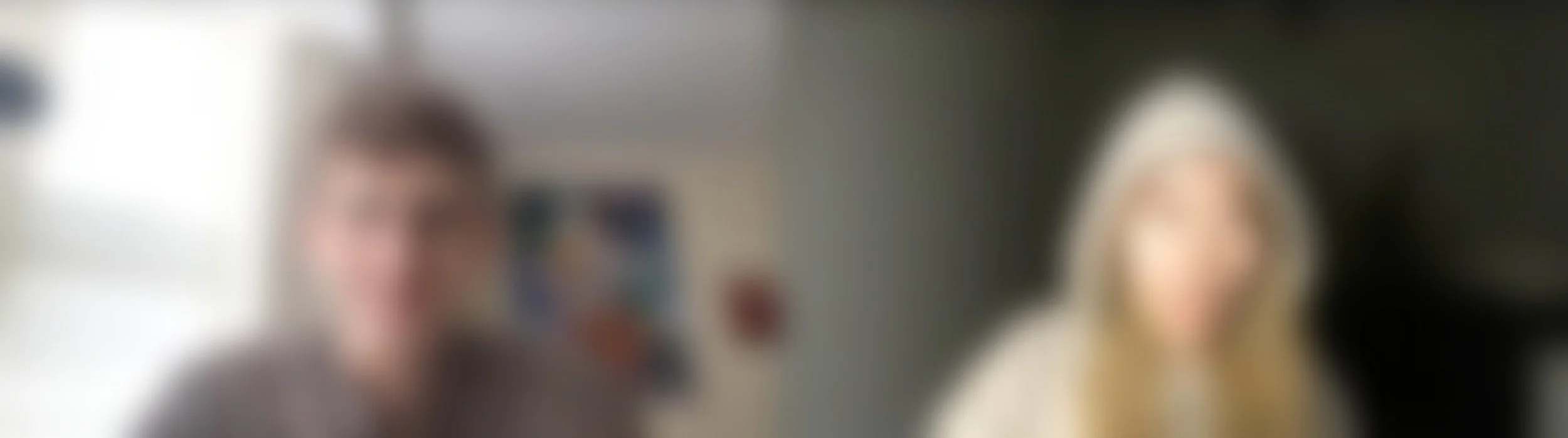 A blurred photo of two people, one male and one female, standing indoors with a neutral background.