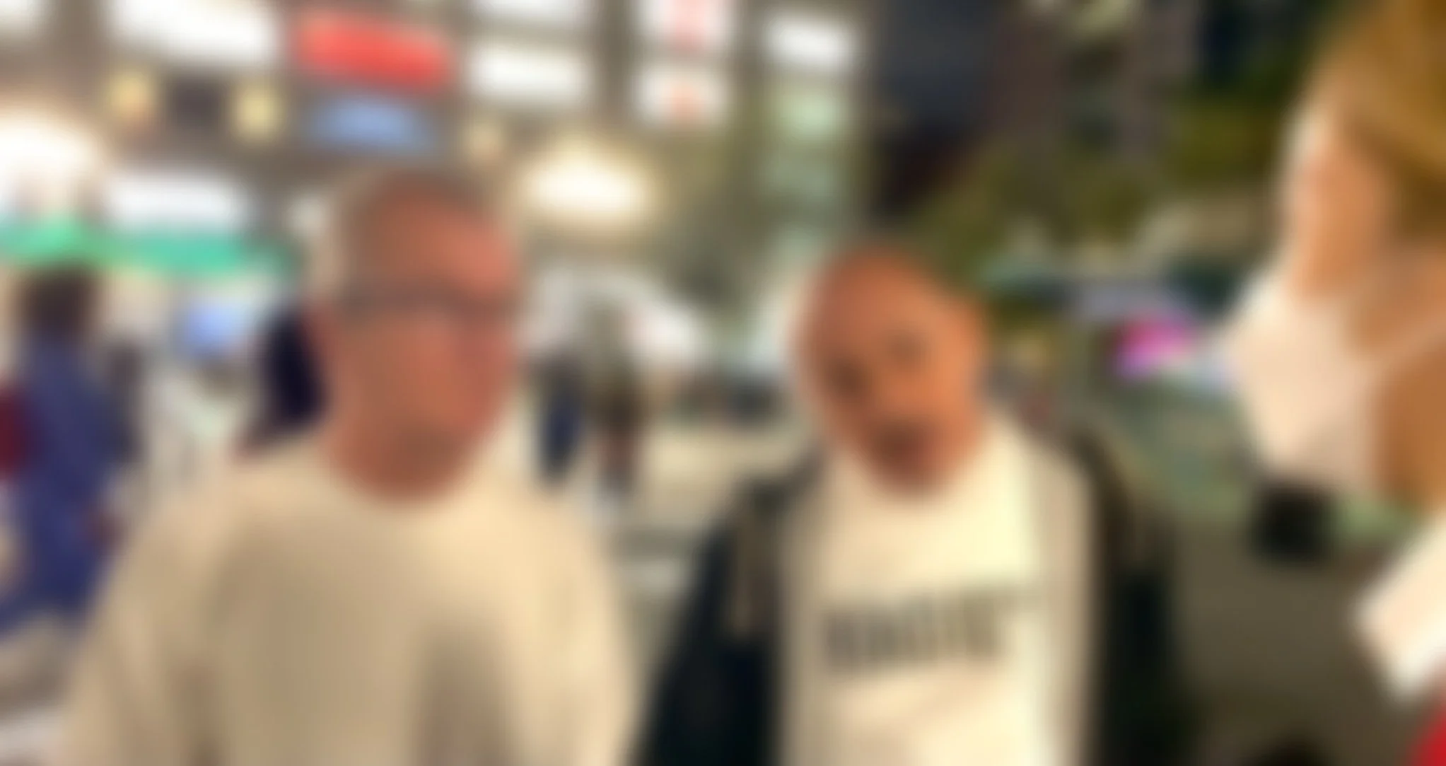 Blurred image of three people standing indoors, likely in a store or restaurant, with shelves and colorful items in the background.