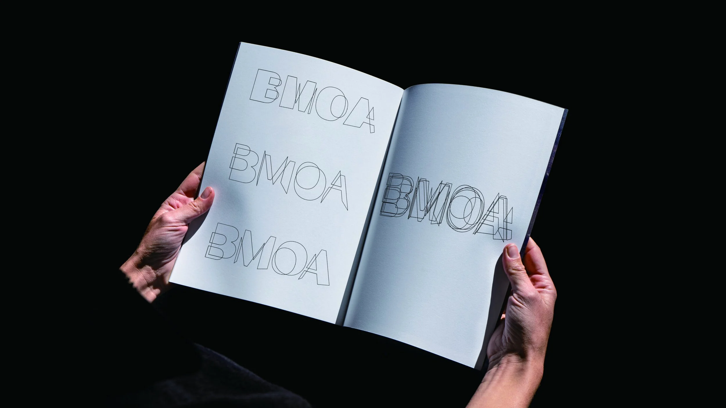 Person holding open sketchbook with repeated outlined text 'BMONA' on white pages in a dark setting.
