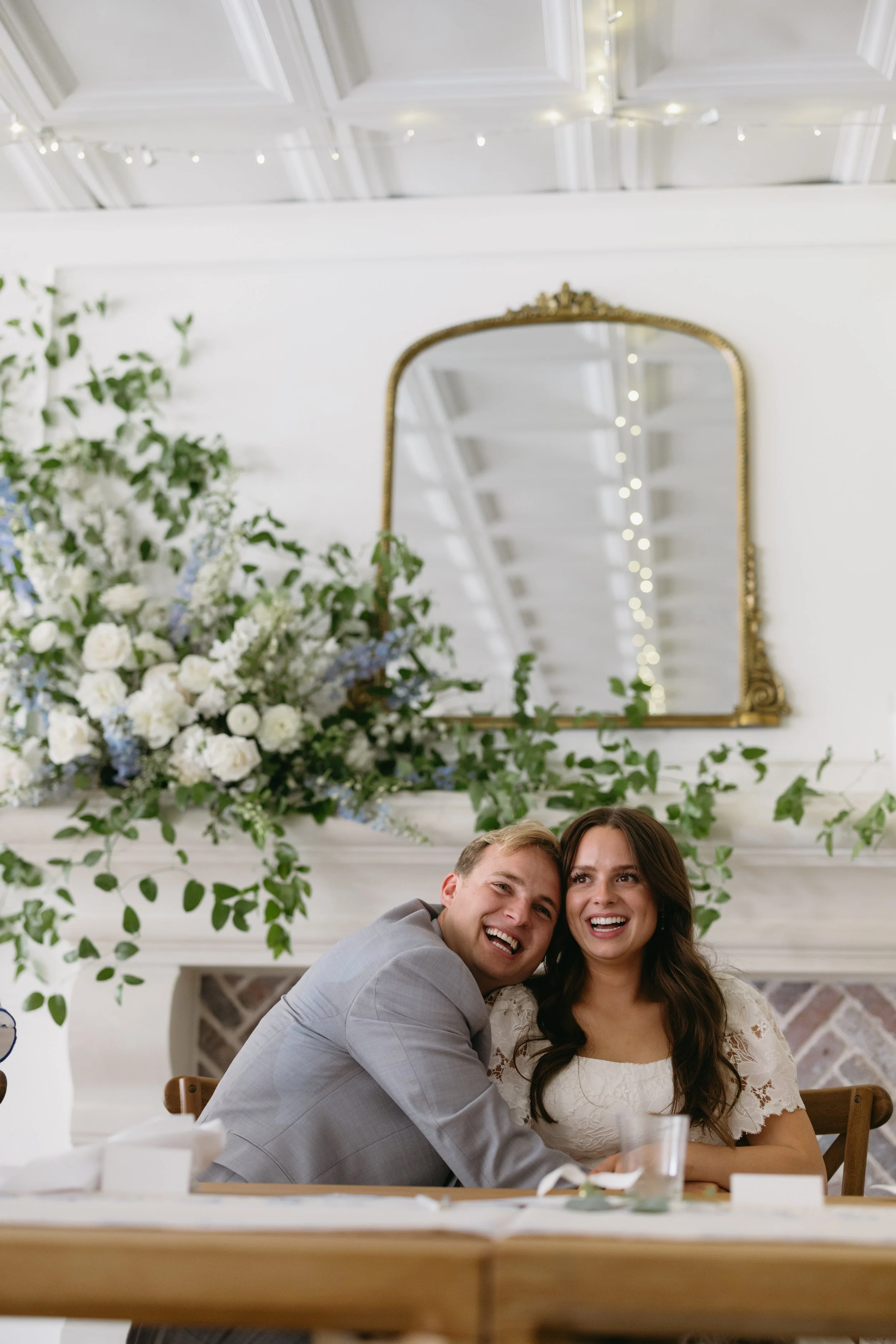 A smiling couple sitting at a table during a celebration, with floral arrangements and a mirror in the background.