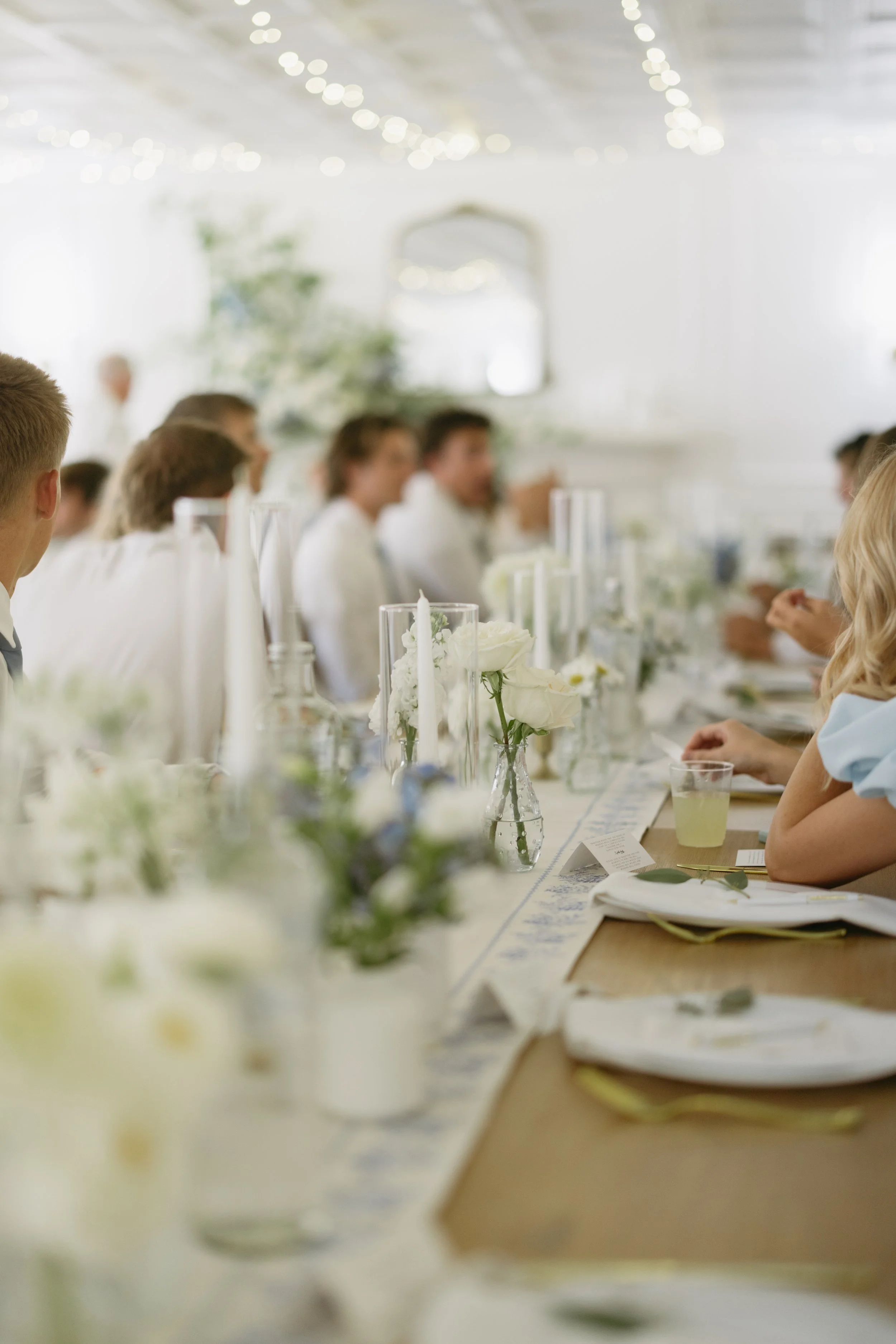 Wedding reception with a long table decorated with white flowers, candles, and table settings, guests seated and engaged in conversation in a bright room with draped ceiling and fairy lights.