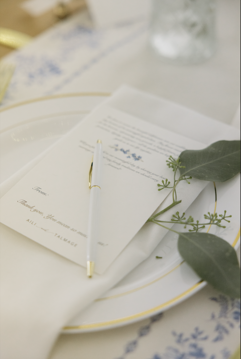 A white pen with gold accents resting on a folded wedding invitation card, decorated with greenery, on a white plate with a gold rim, placed on a table with a patterned cloth.