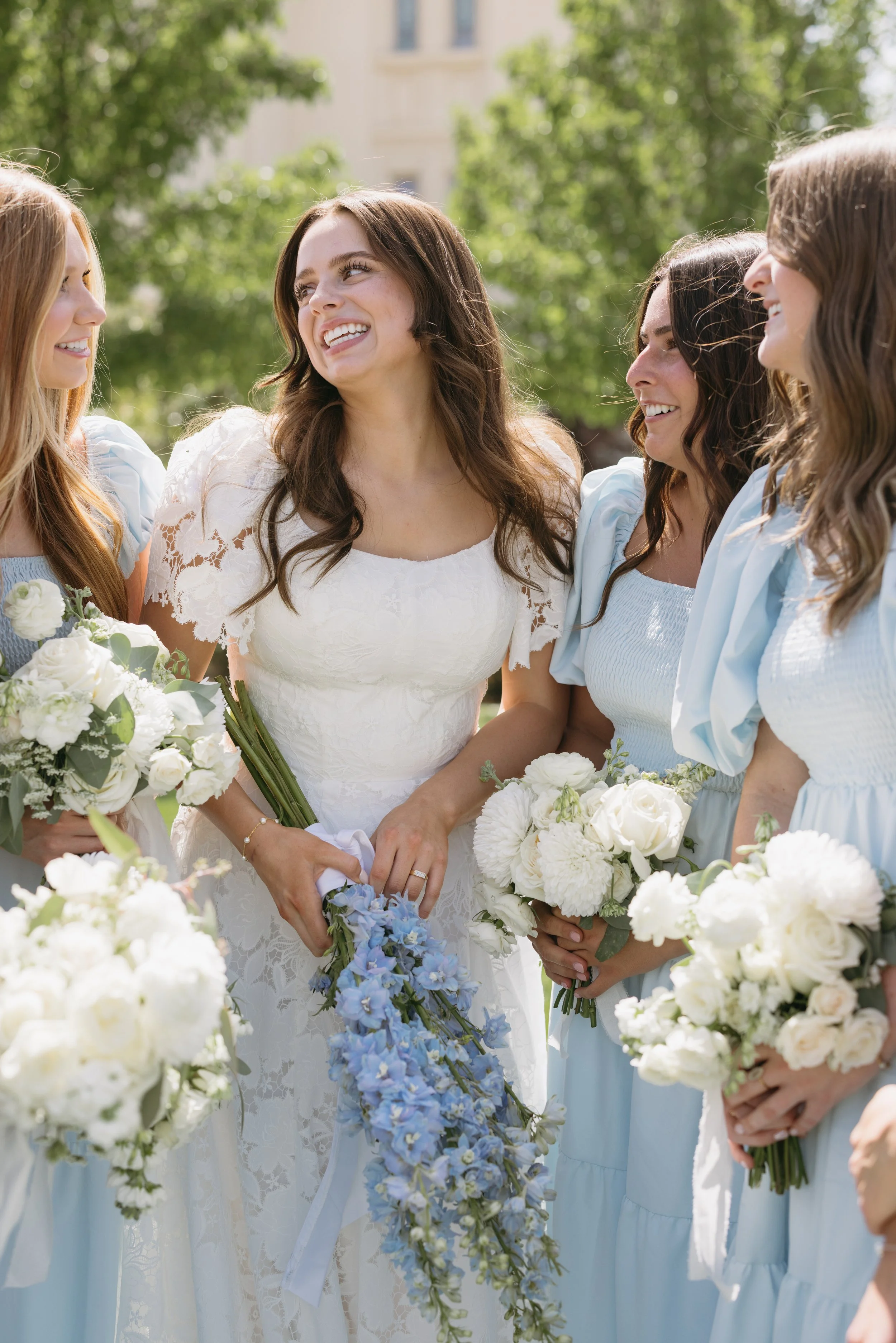 Group of women in wedding dresses holding bouquets, smiling and talking outdoors during a wedding celebration.