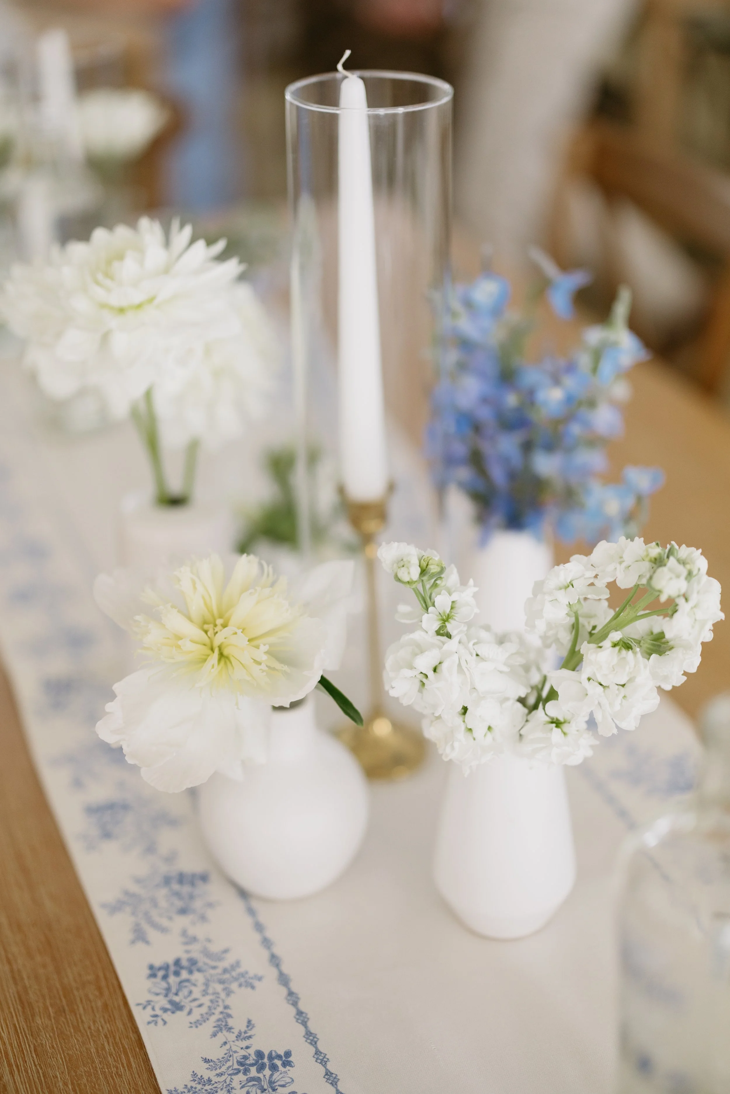 A table decorated with white and blue flowers in white vases, a tall glass candleholder with a white taper candle, and a patterned table runner.