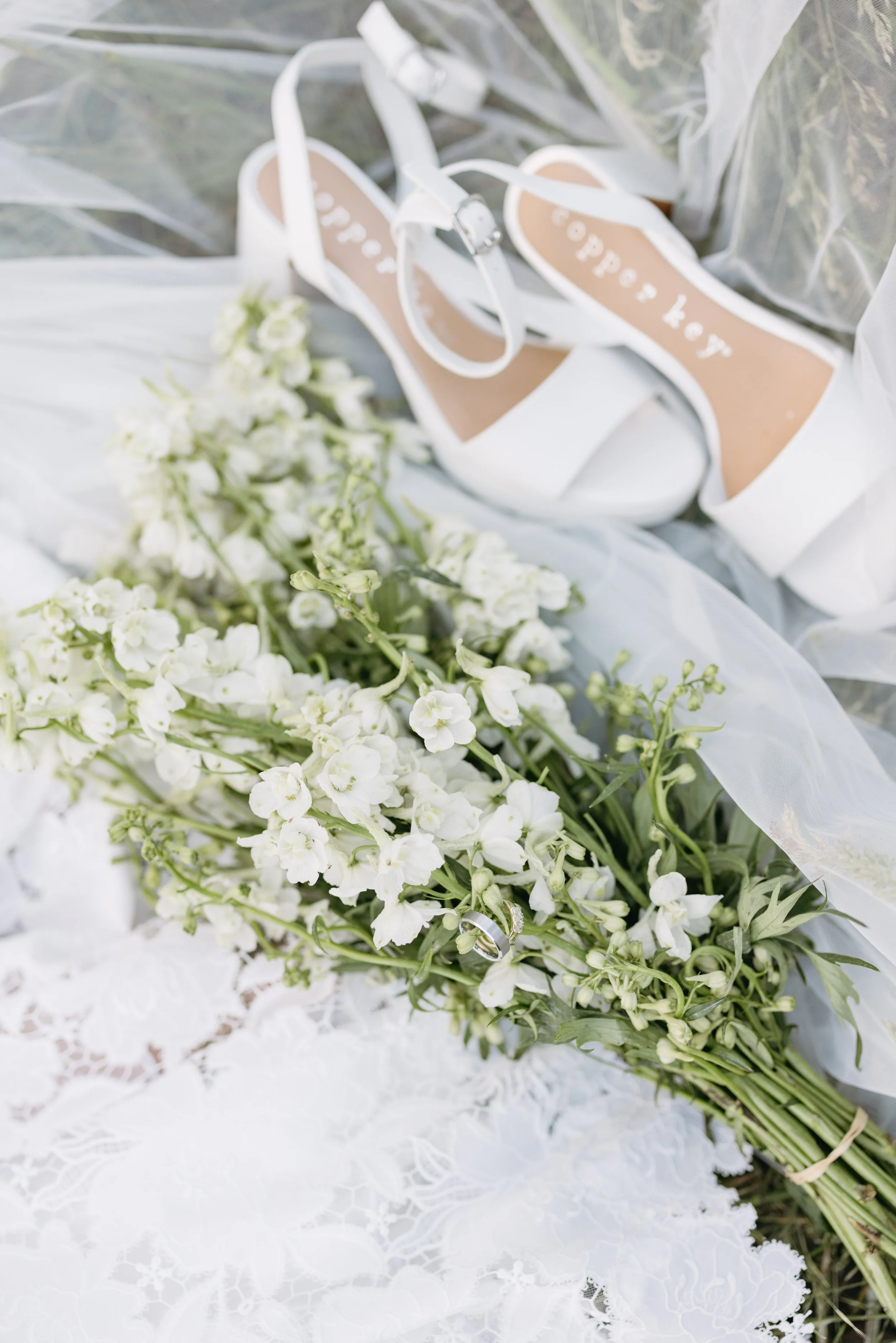 White high-heeled shoes with ankle straps and the words "happily" written inside, a bouquet of white flowers, and a silver ring, all arranged on white lace fabric.