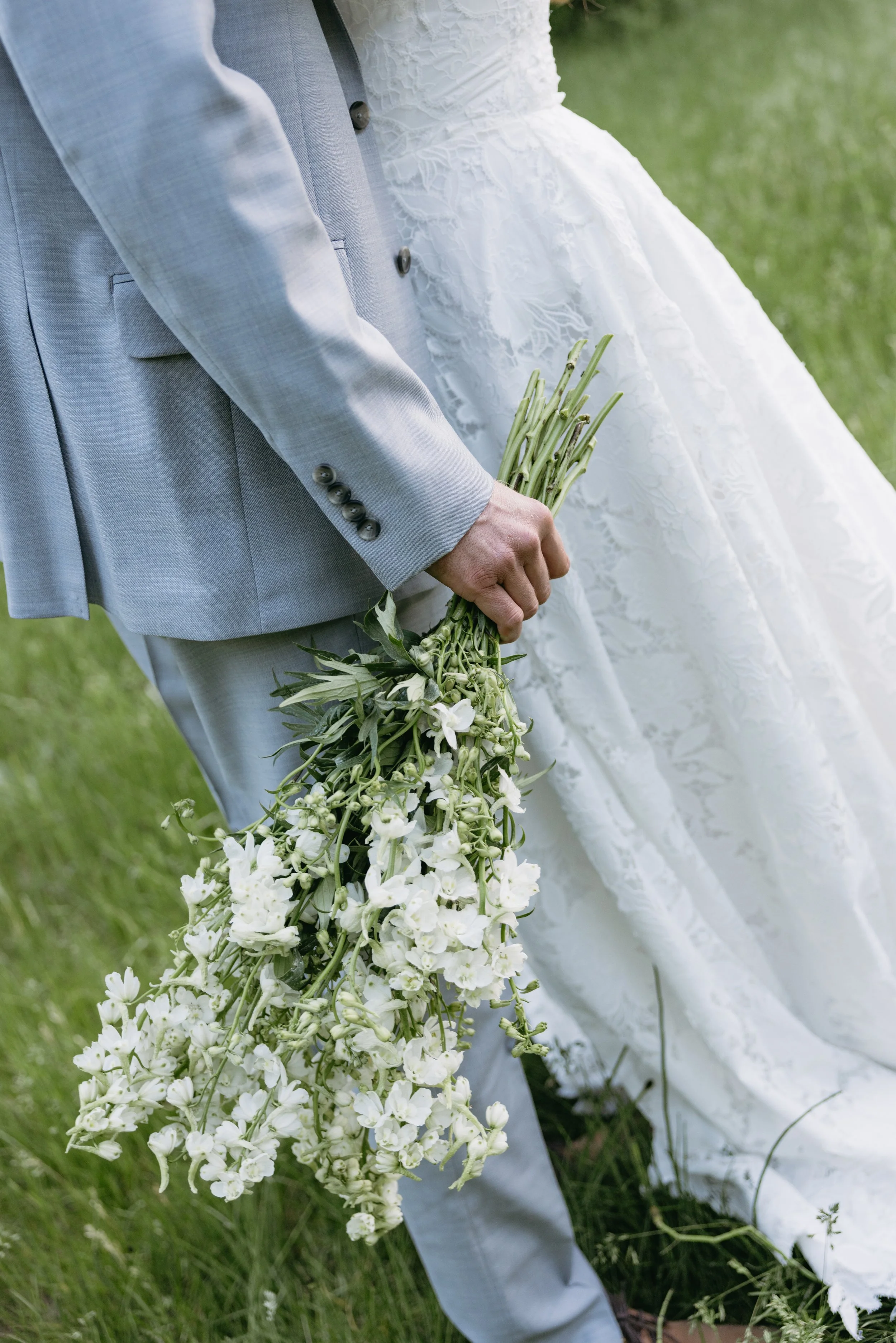 A groom in a light gray suit holding a large bunch of white flowers outdoors.