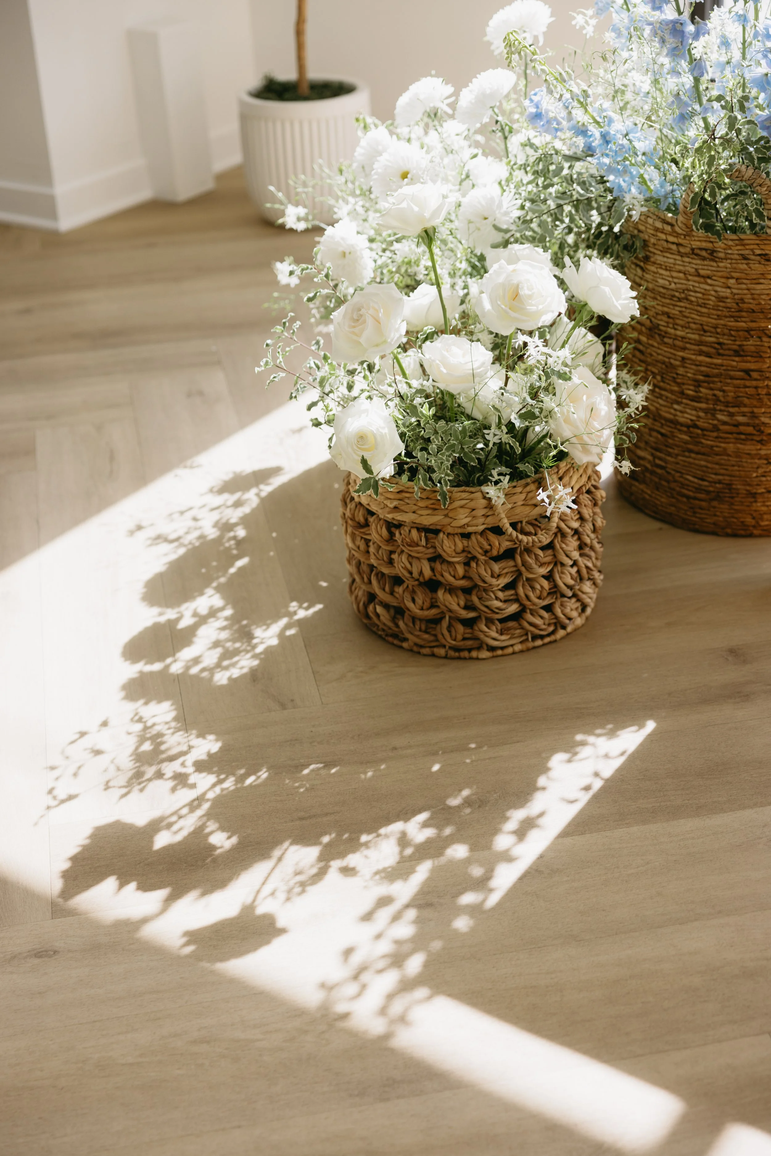 A woven basket filled with white roses and greenery, casting shadows on a light wooden floor in a well-lit room.