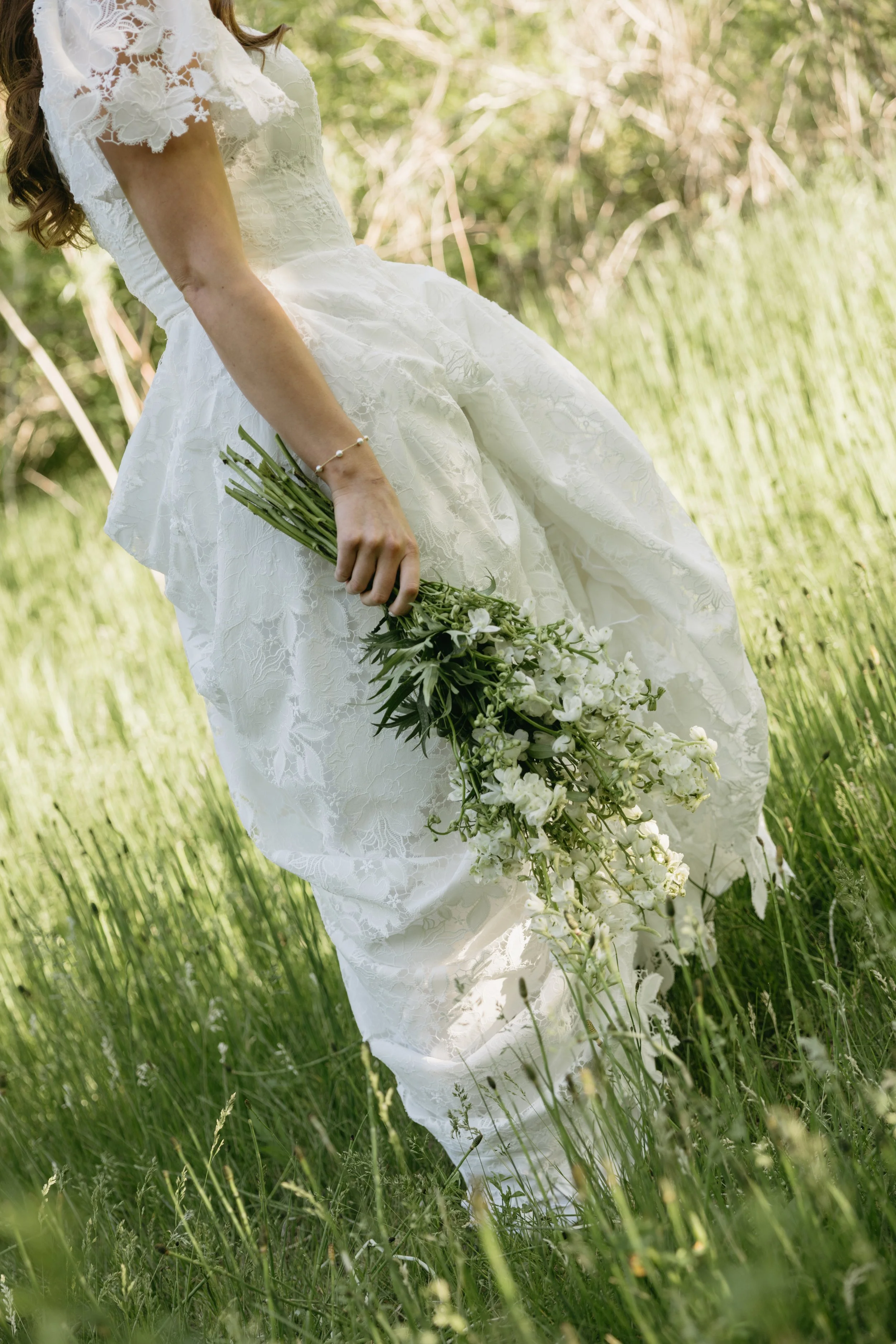 A woman in a white lace wedding dress holding a bouquet of white flowers while standing in tall grass outdoors.