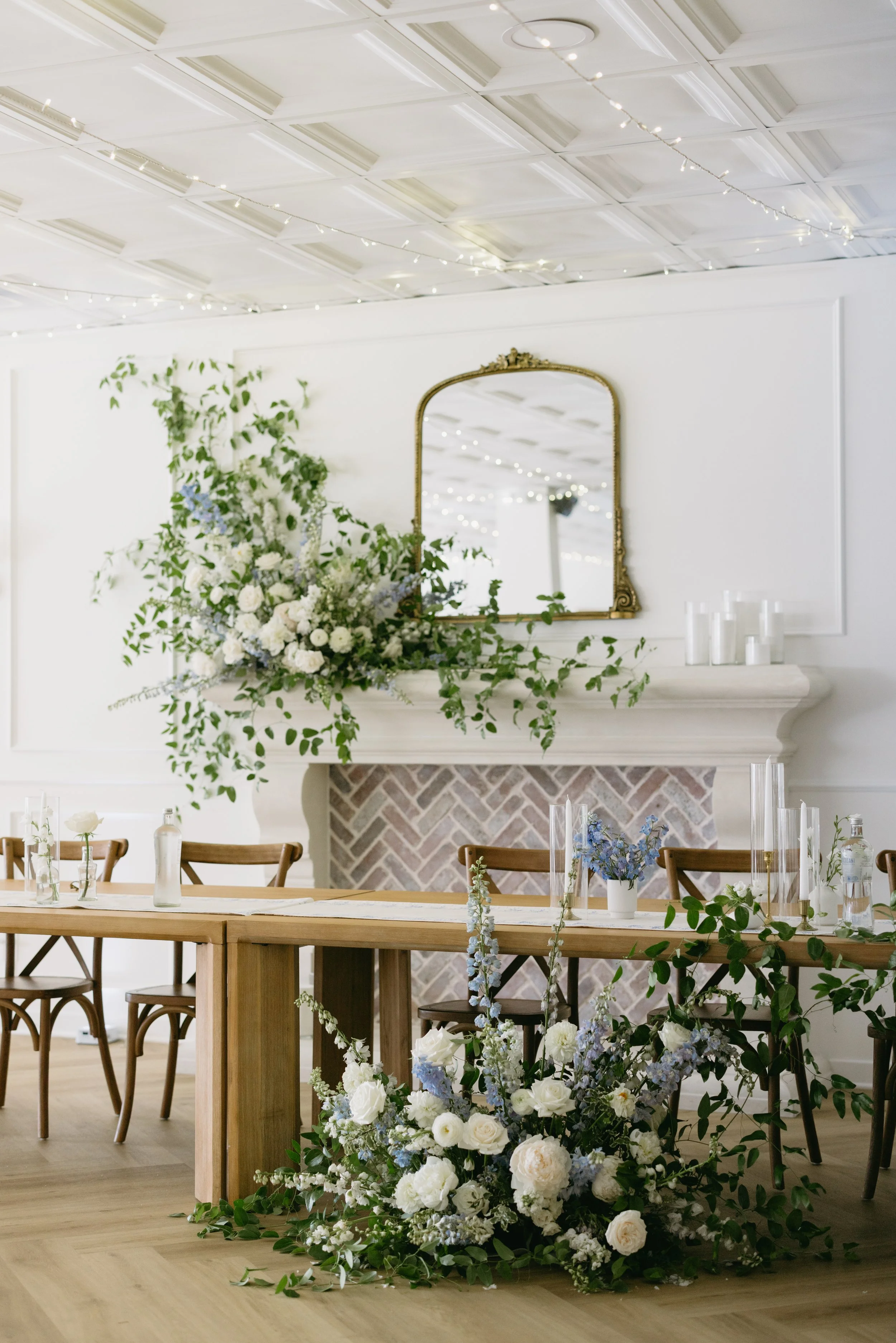 Elegant indoor event space with a long wooden table decorated with white and blue flowers, candles, and greenery. A large flower arrangement is on the fireplace mantle, and a mirror and string lights adorn the white paneled ceiling.