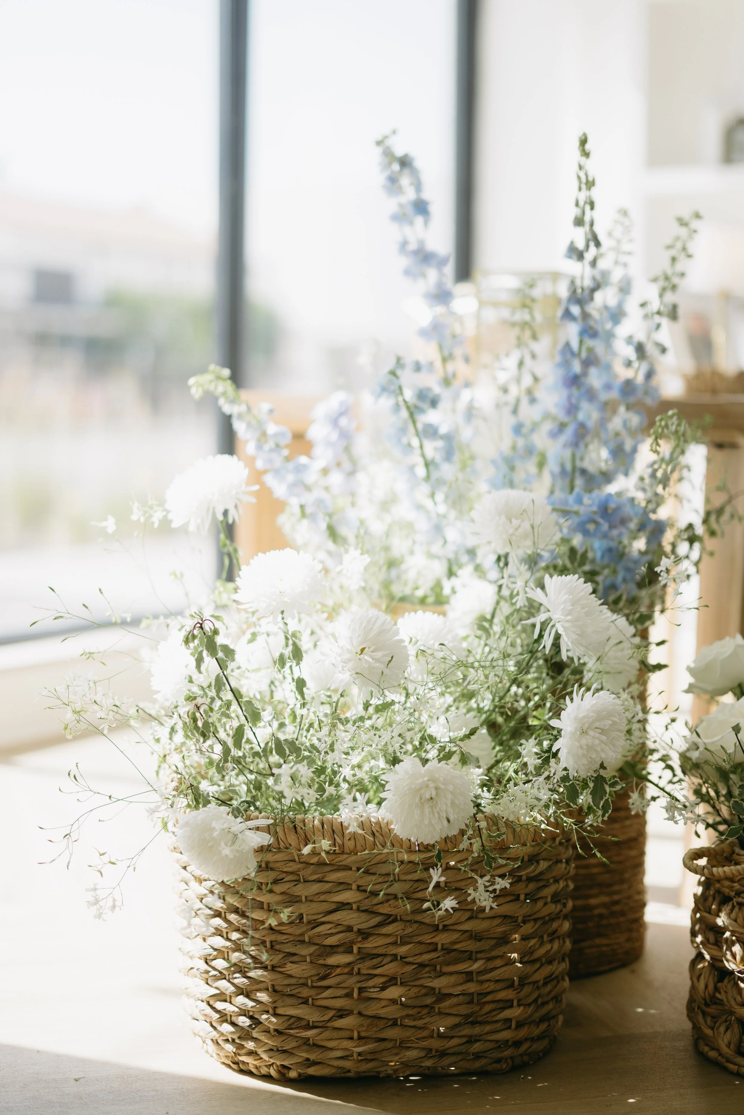 A woven basket filled with white and blue flowers, placed near a window with sunlight coming through.