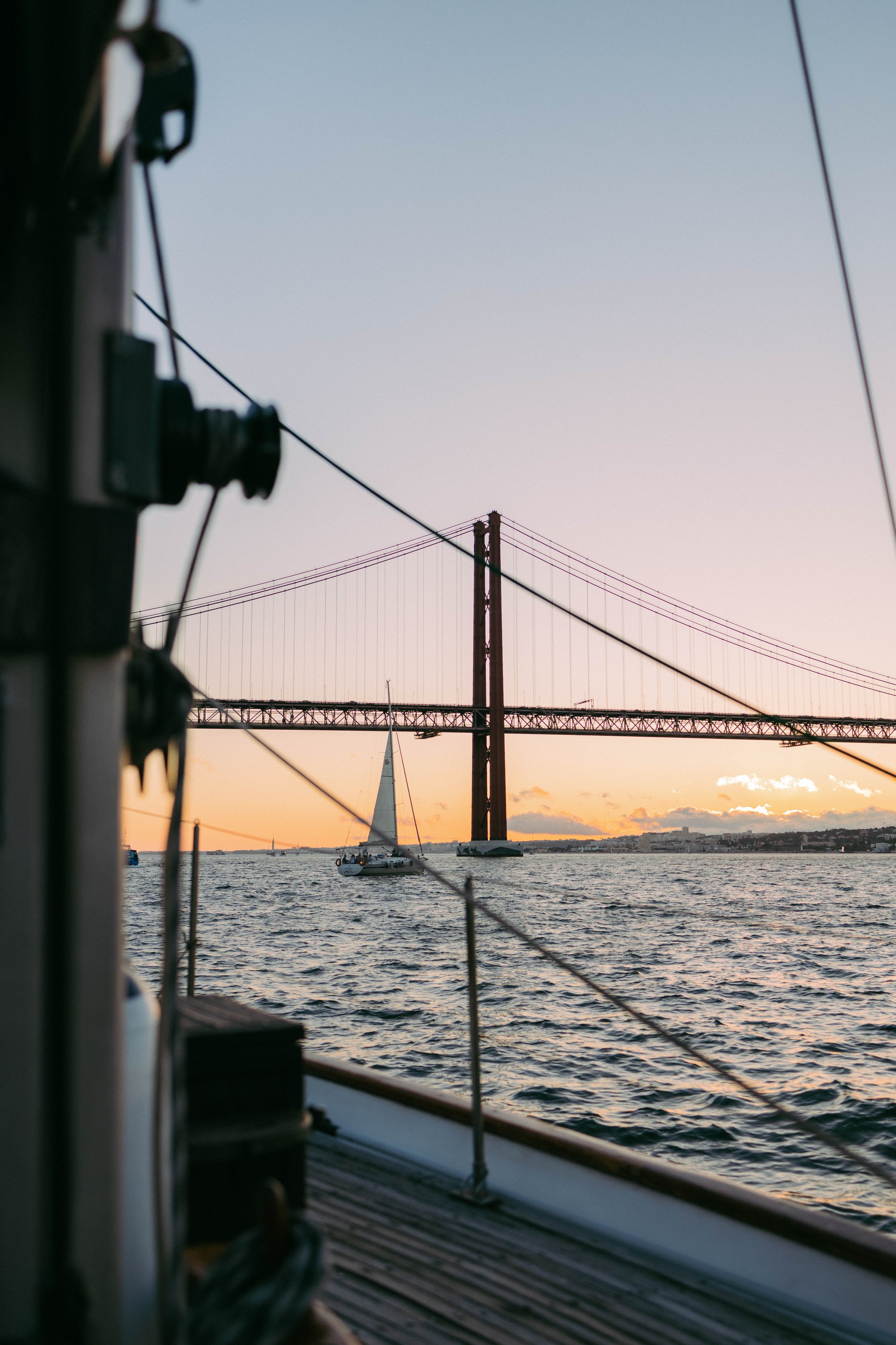 View of a sailboat sailing under a bridge at sunset, seen from the deck of another boat.