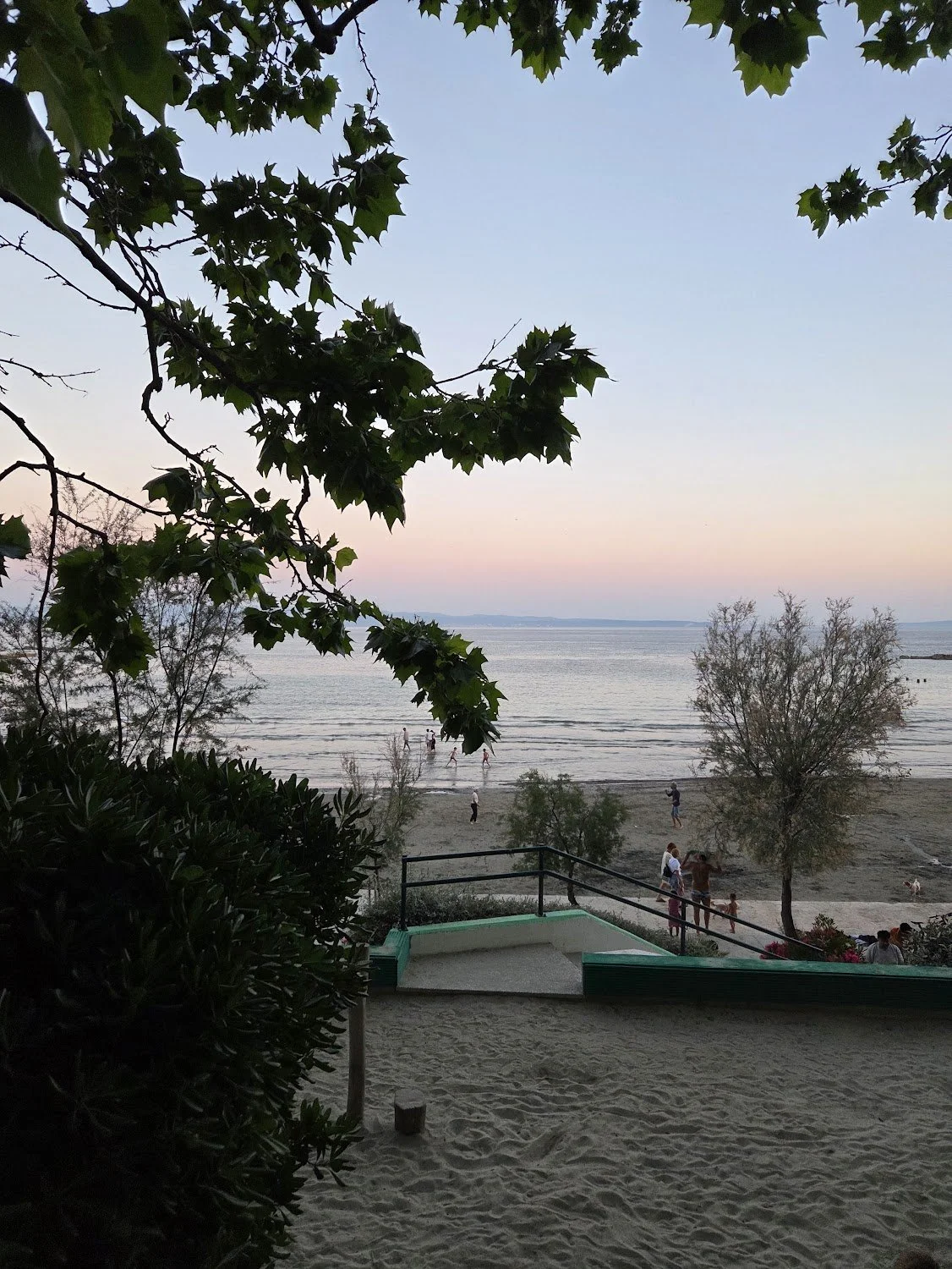 View of a beach scene at sunset with trees in the foreground, calm water, and people enjoying the shoreline.