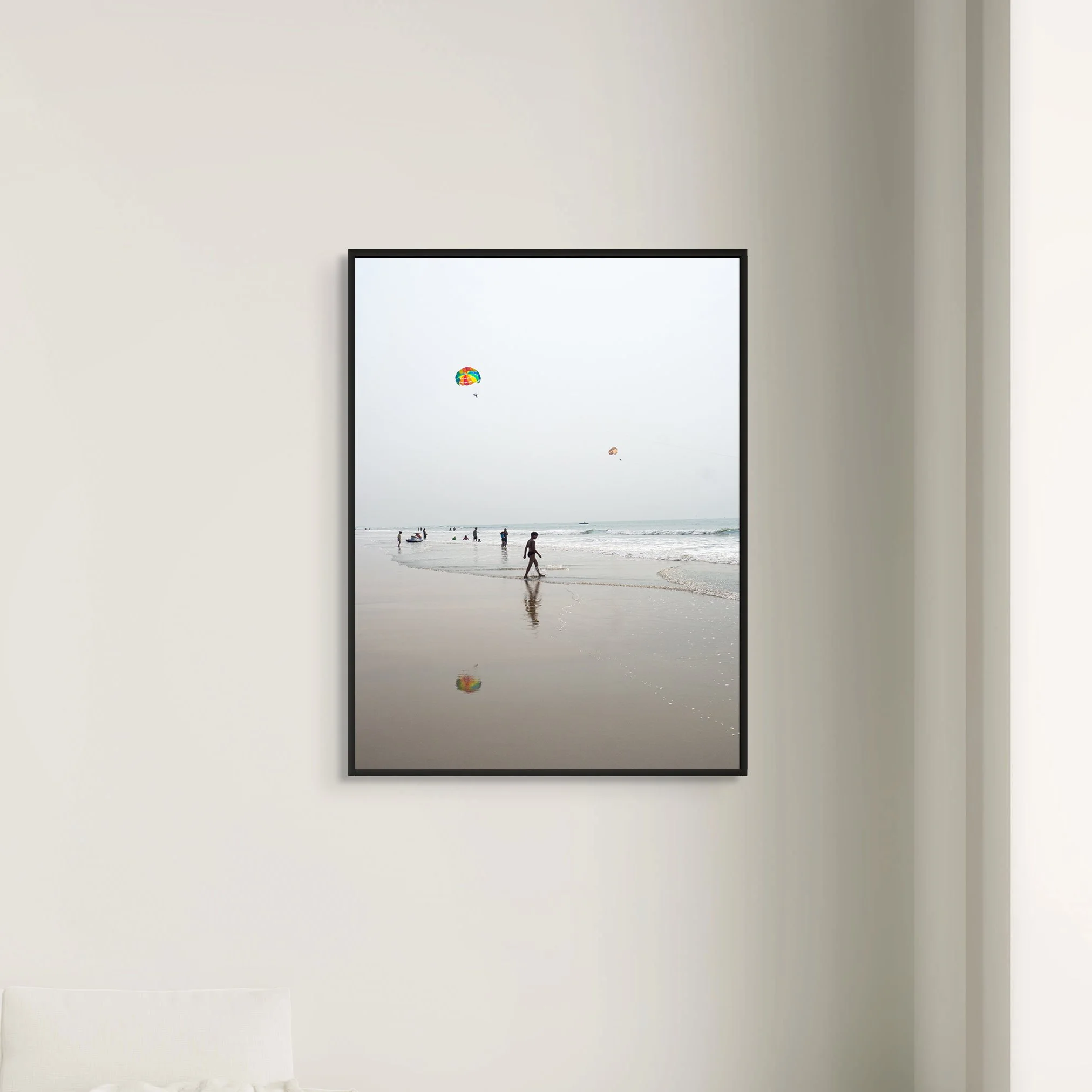 The image shows a vertical framed mirror on a plain light-colored wall. The mirror reflects a beach scene with people, umbrellas, and boats in the water.