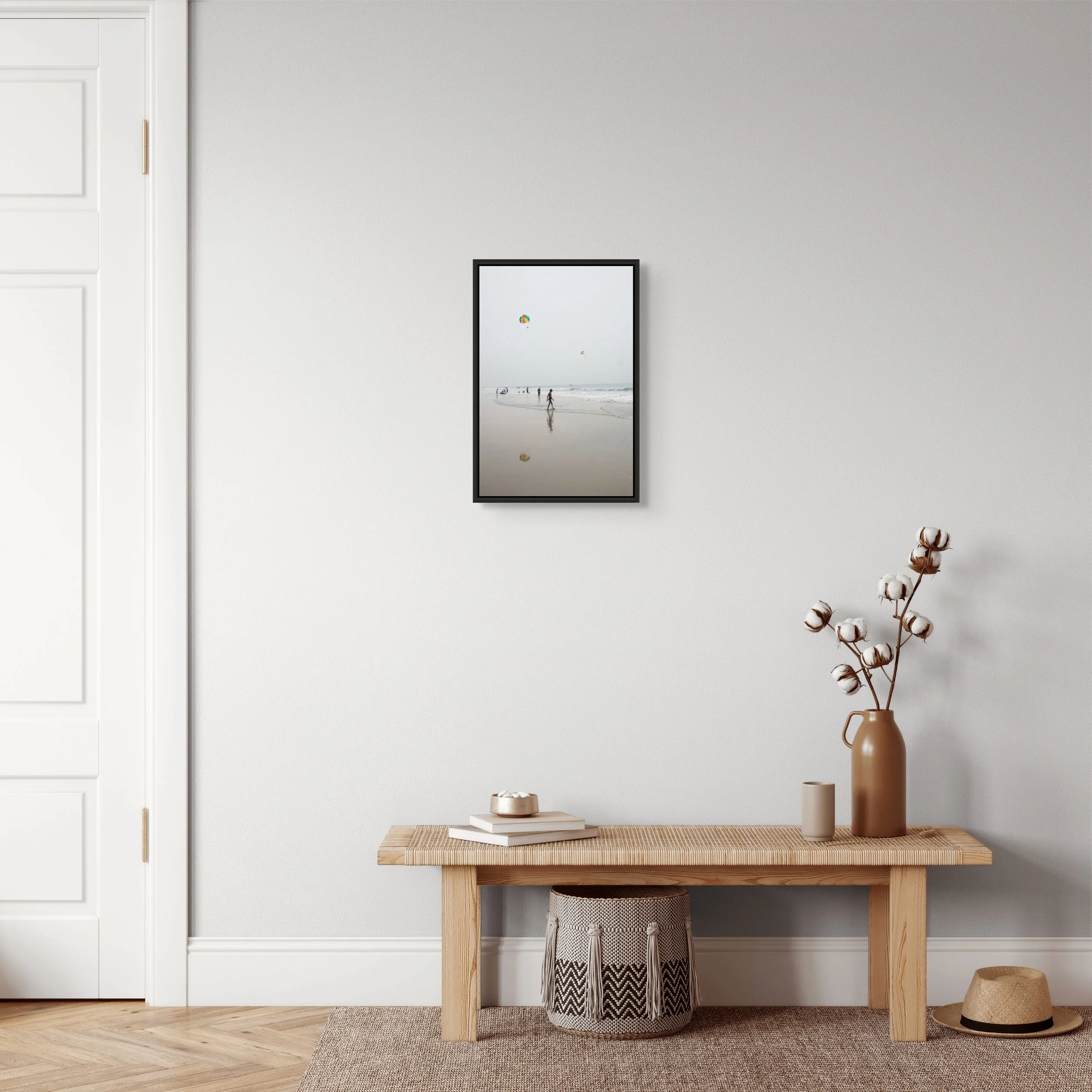 Interior scene with a light-colored wall, a framed beach photo, a wooden table, and a tall beige vase with cotton stems.