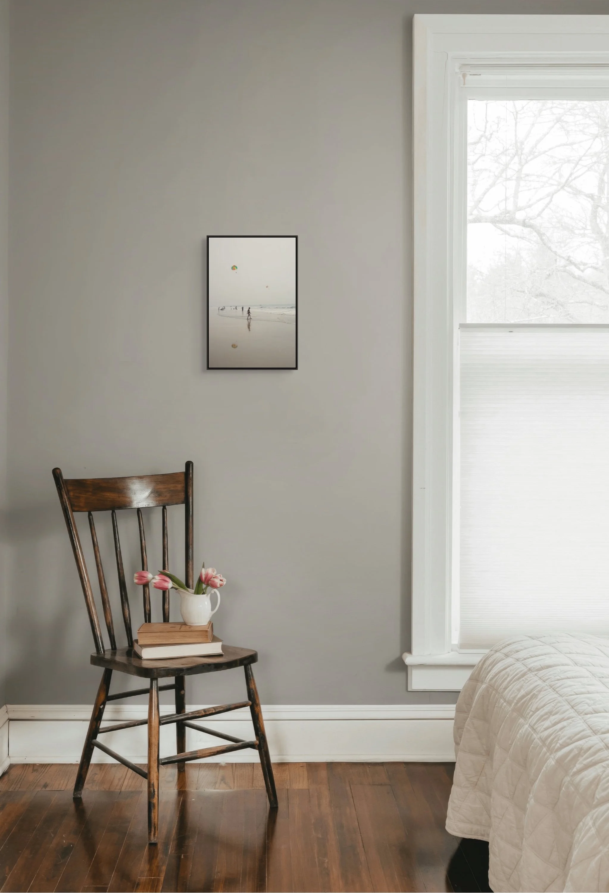 A cozy bedroom corner with a wooden chair holding a white vase with pink tulips. The chair has a stack of books. To the right is a bright window with white trim, and a wall with gray paint and a framed beach photograph. Part of a bed with a beige qui