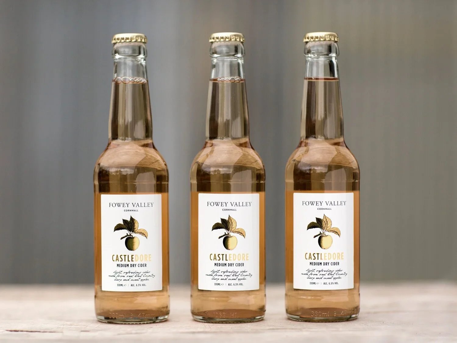 Three bottles of Fowey Valley Castle Dore medium dry cider on a wooden surface against a neutral background.