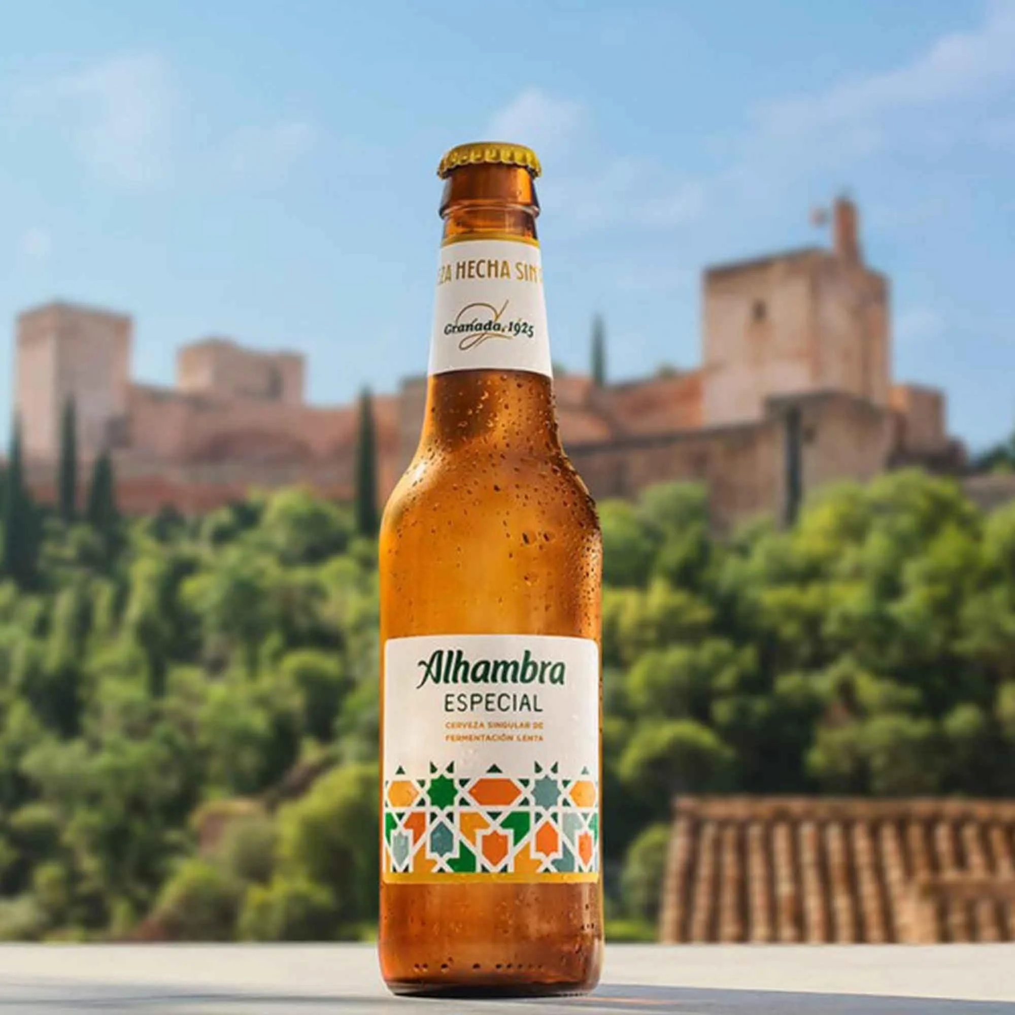 A bottle of Alhambra Especial beer with condensation, set against a blurred background of a historic Spanish castle on a hill and green trees.
