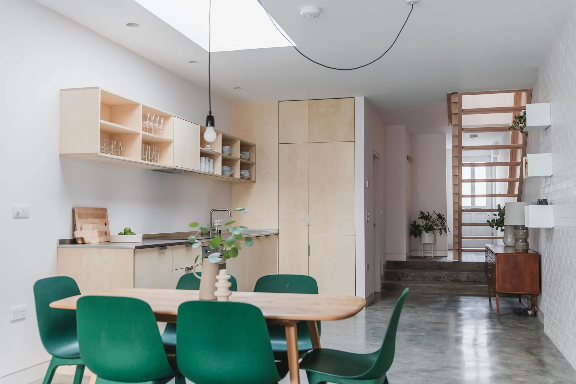 Marjoram Architects — Modern kitchen and dining area with wooden table and green chairs, white cabinets, open shelving with glassware and dishes, wooden staircase, and pott