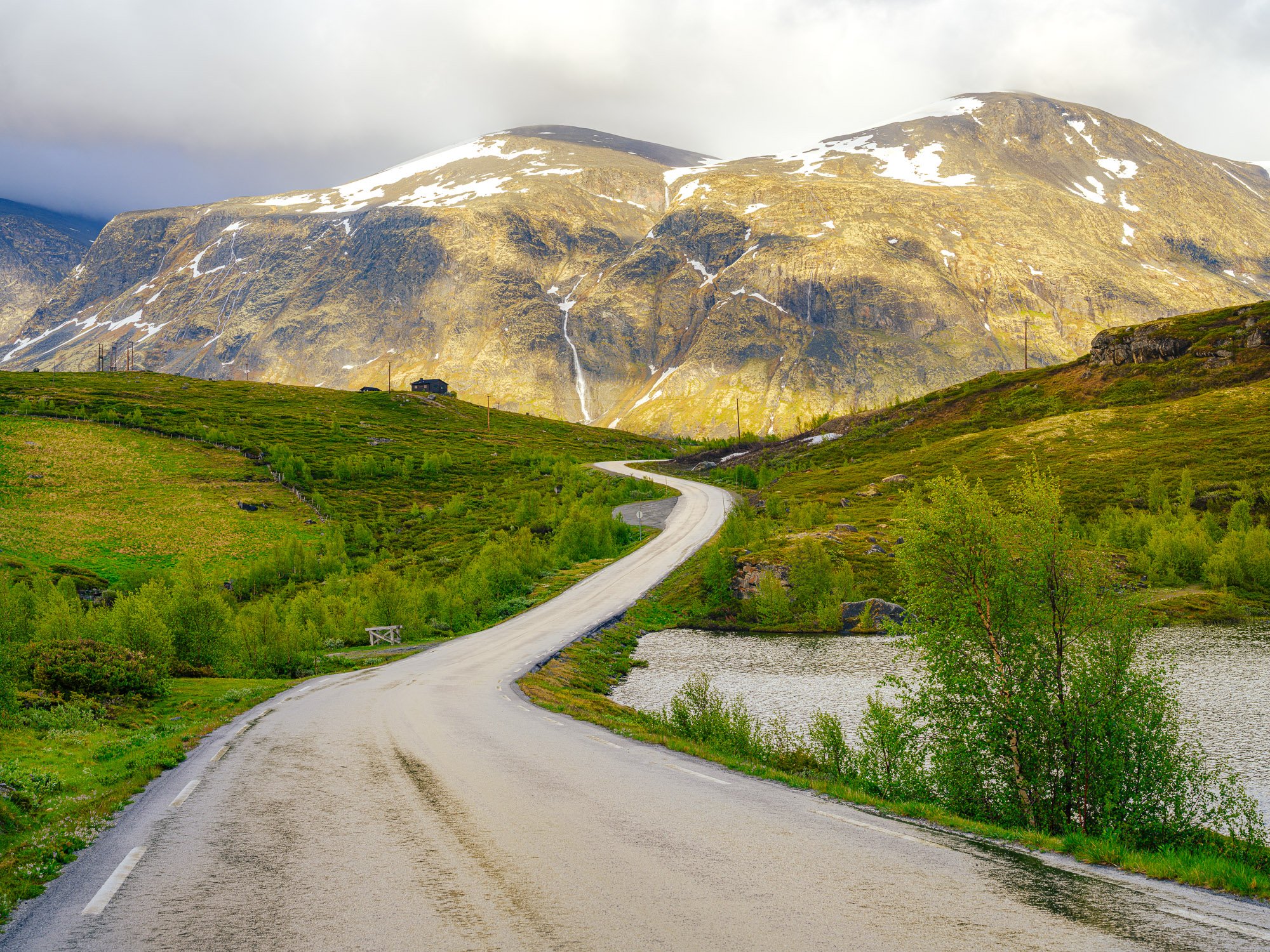 En svingete vei som går gjennom grønt landskap med innsjøer, omgitt av fjell med snø på toppen, under en delvis overskyet himmel.